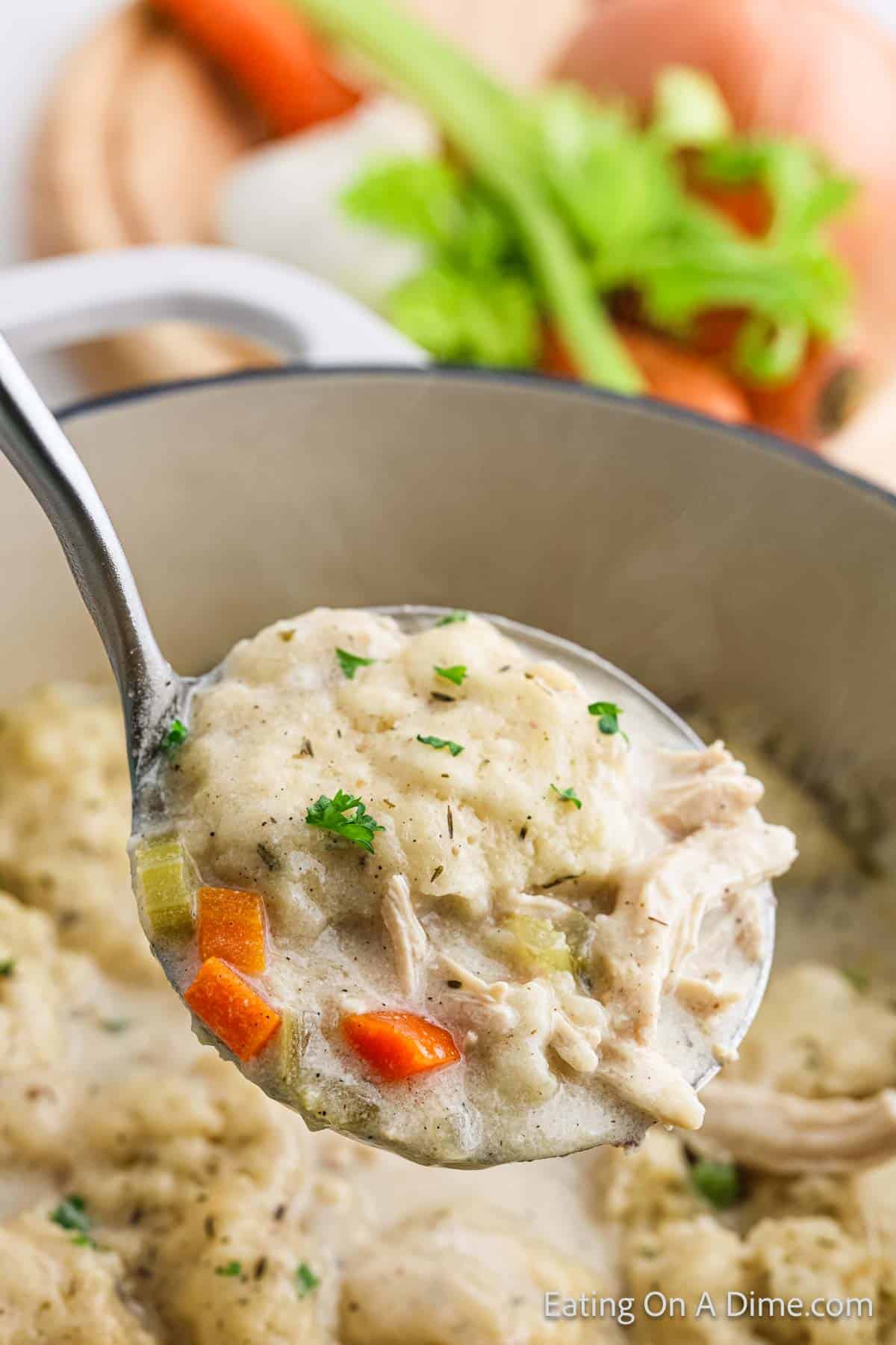 A ladle brims with homemade chicken and dumplings, showcasing tender chunks of chicken alongside diced carrots and celery in a creamy broth. Fresh parsley elegantly garnishes this classic dish, while a blurred cutting board hints at the fresh ingredients used to create this comforting meal.