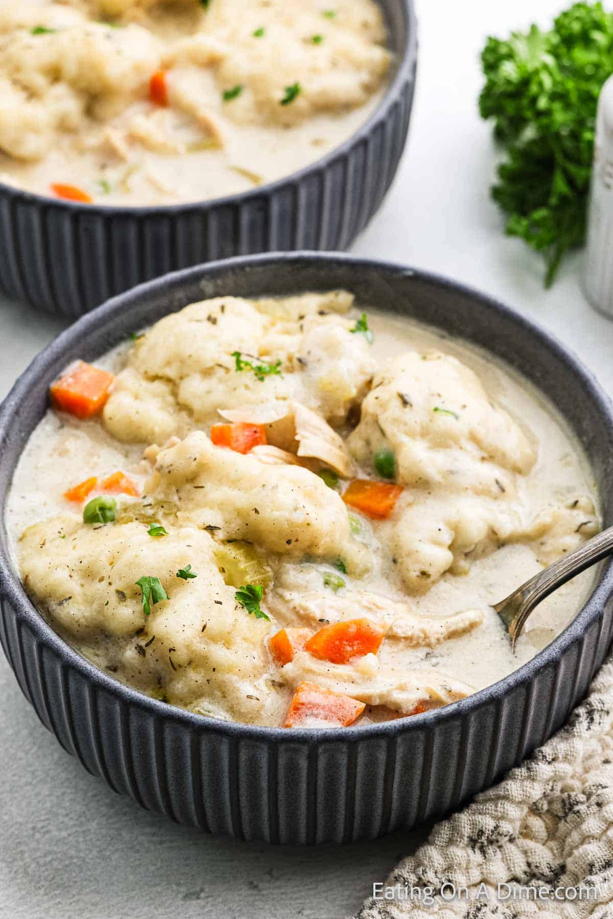 A bowl of chicken and dumplings with creamy broth, showcasing fluffy dumplings and chunks of carrots. A second bowl is partially visible in the background, topped with a garnish of fresh herbs.