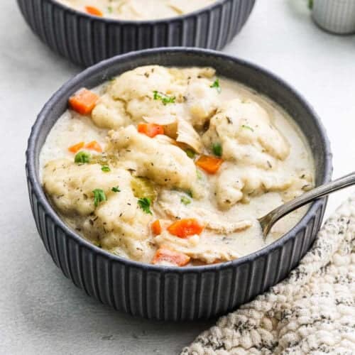 A bowl of creamy chicken and dumplings, garnished with chopped parsley, features tender chicken pieces with chunks of carrots and celery. A spoon rests invitingly in the bowl, set on a textured cloth, while another dish tantalizingly peeks from the background.