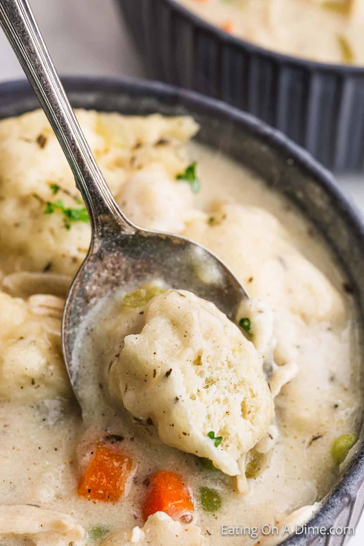 A bowl of creamy chicken and dumplings with tender pieces of carrot and celery. A spoon lifts a fluffy chicken dumpling from the bowl, showcasing its delightful texture. Another bowl peeks from the background, hinting at seconds.