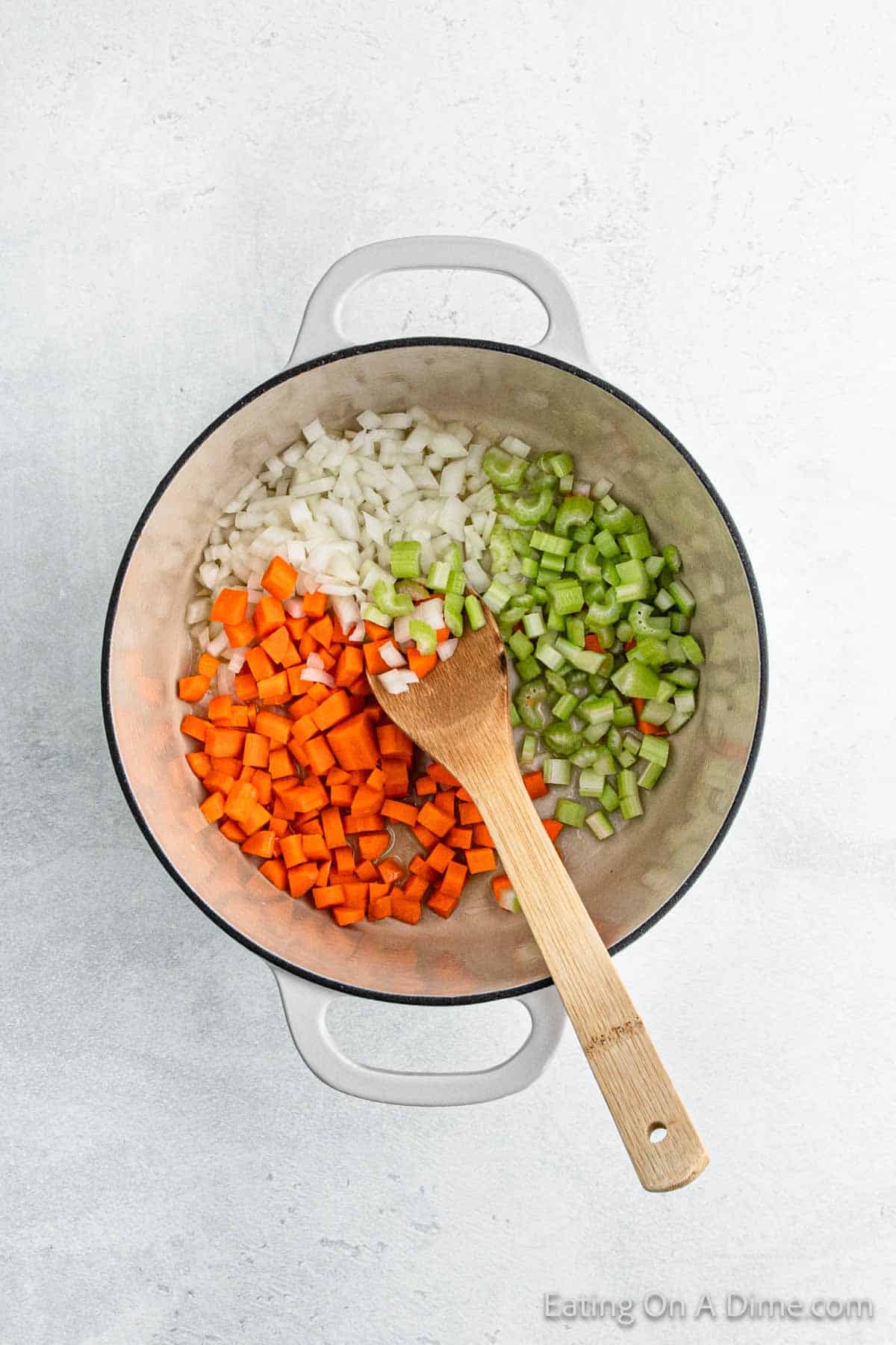 A pot with diced onions, carrots, and celery sits ready for a hearty chicken stew. A wooden spoon rests inside, hinting at the delicious addition of savory dumplings soon to come. The colorful arrangement is set against a light, textured background, promising comfort in every bite.