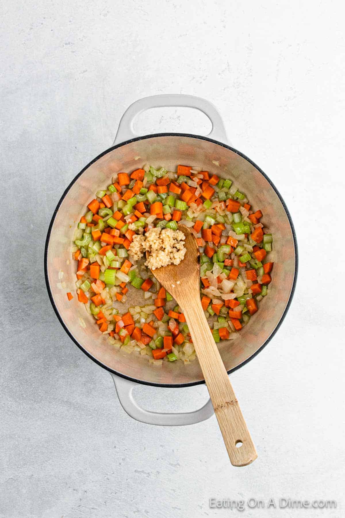 A pot containing chopped carrots, celery, and onions sautés with a wooden spoon resting inside. The mixture starts to brown as tender chicken pieces join the medley, simmering away. Imagine fluffy dumplings soon completing this hearty dish on a light-colored surface.