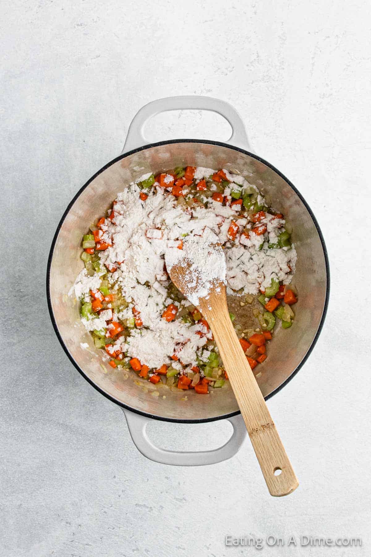 A white pot on a light surface contains chopped carrots, celery, and onions topped with flour, hinting at a comforting chicken dumpling dish. A wooden spoon rests inside, partially covered in flour, as the ingredients are mixed to begin this hearty recipe preparation.