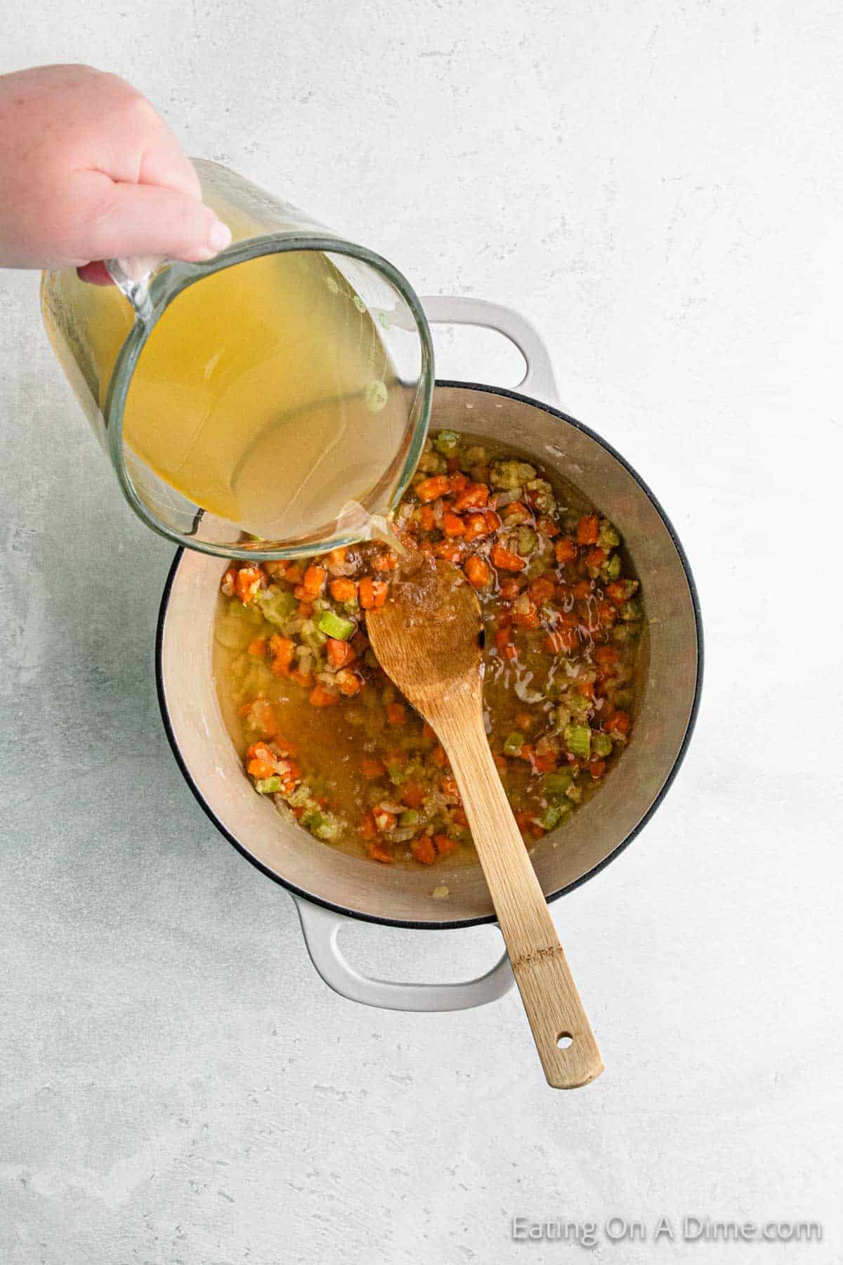 A hand pours broth from a glass pitcher into a pot filled with chopped carrots, celery, and onions for a comforting chicken recipe. A wooden spoon rests in the pot, which is set against a light-colored surface.