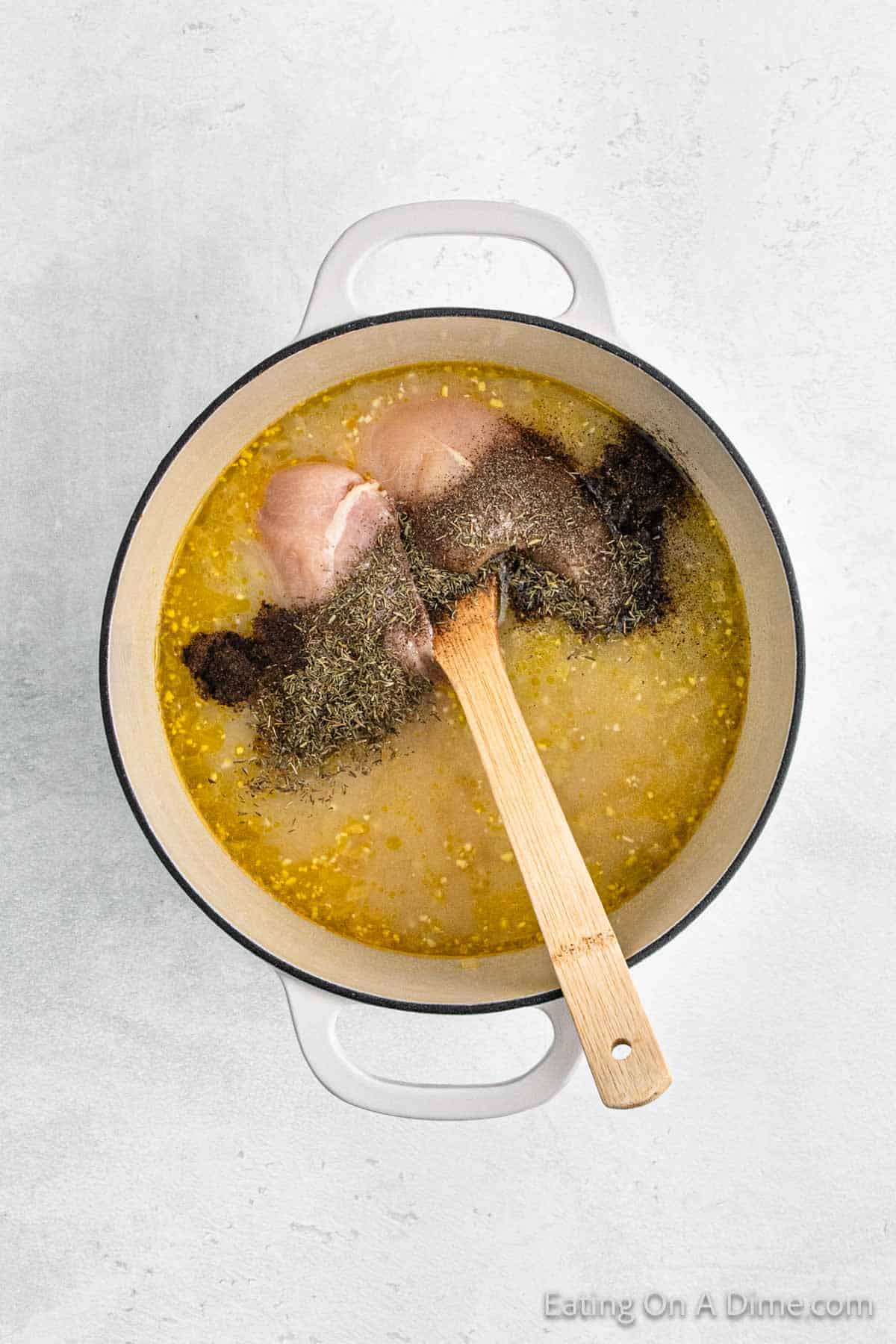 A pot filled with chicken breast, broth, and seasonings, accompanied by dumplings ready to simmer. A wooden spoon rests on the side as this delicious mixture begins its journey on a light-colored countertop.