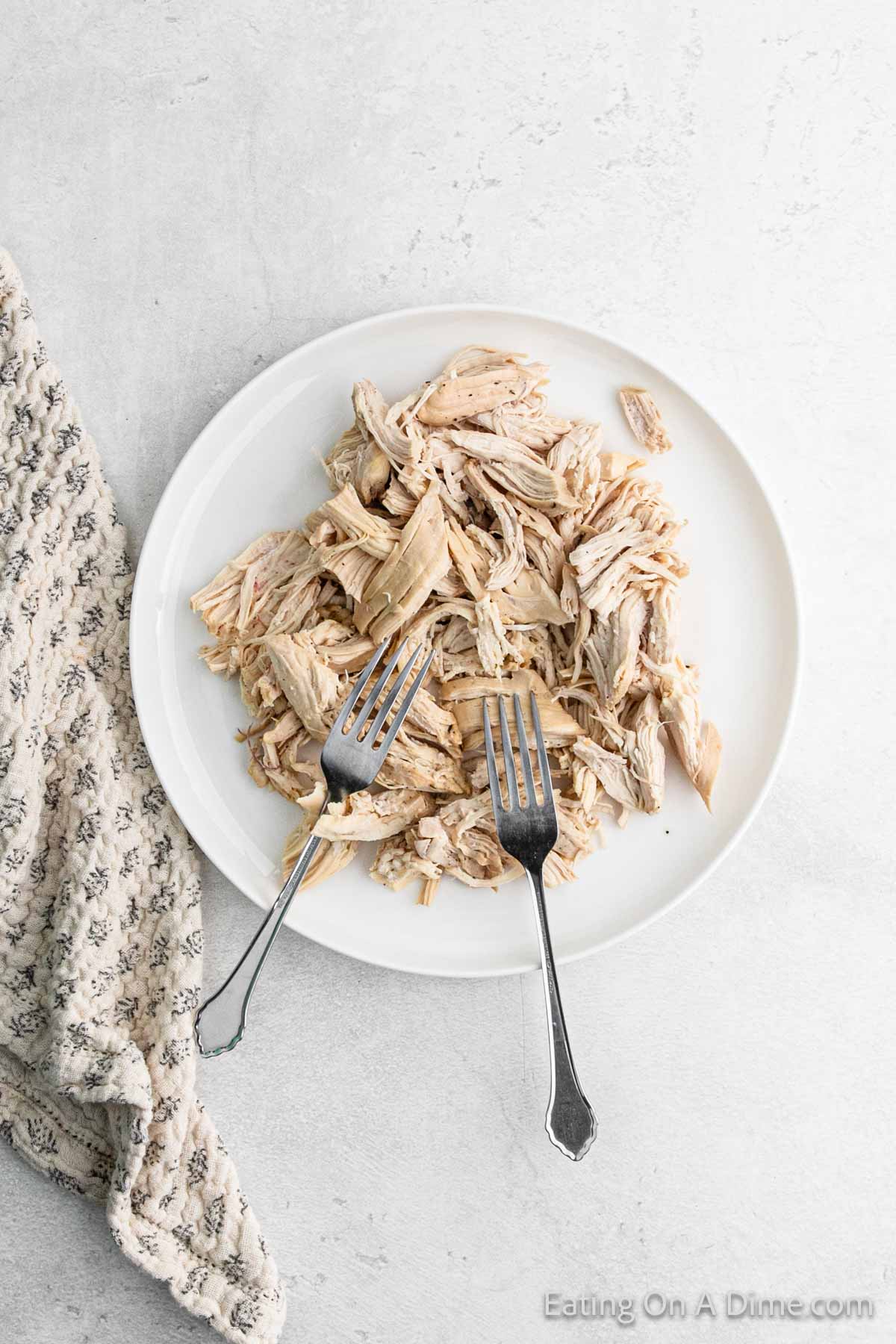 A white plate with shredded chicken sits alongside two forks on a textured white surface, perfect for trying a new recipe. A patterned cloth napkin is placed beside the plate, ready to serve your delicious creation.