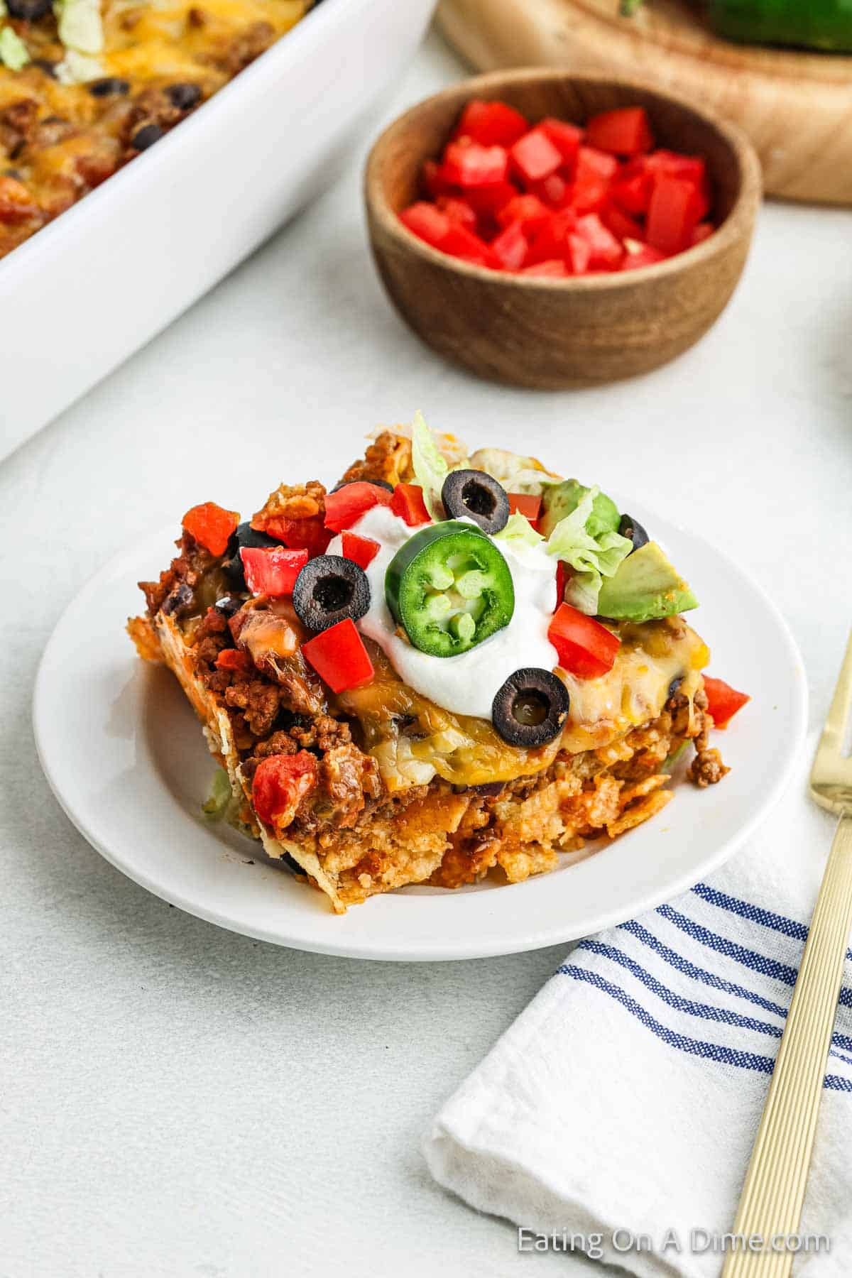 A serving of taco casserole on a plate topped with slice jalapenos, sour cream, slice black olives and diced tomatoes