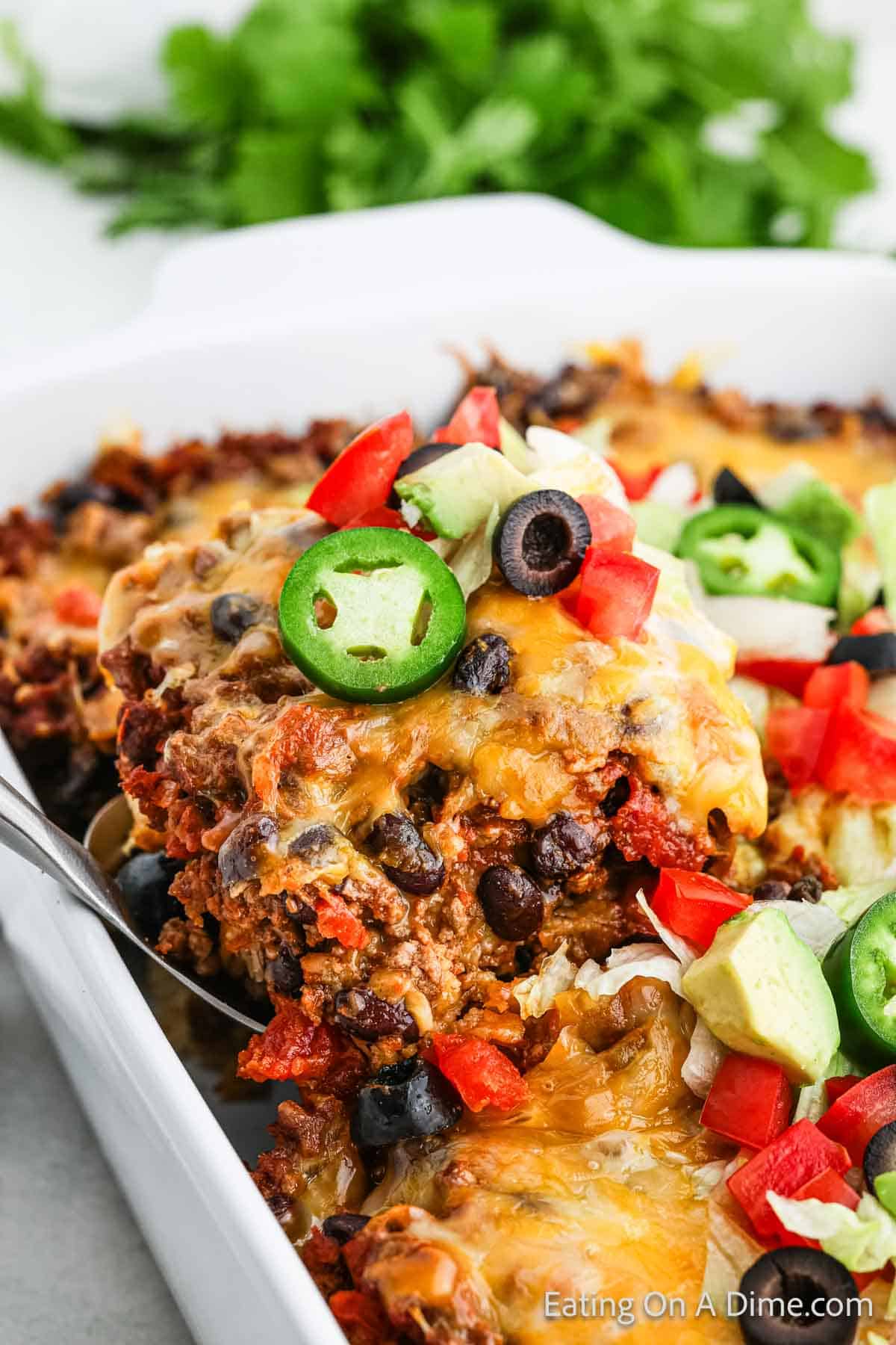 Taco Casserole in a baking dish topped with melted cheese, slice jalapeno peppers, black olives, diced tomatoes with a serving on a spatula