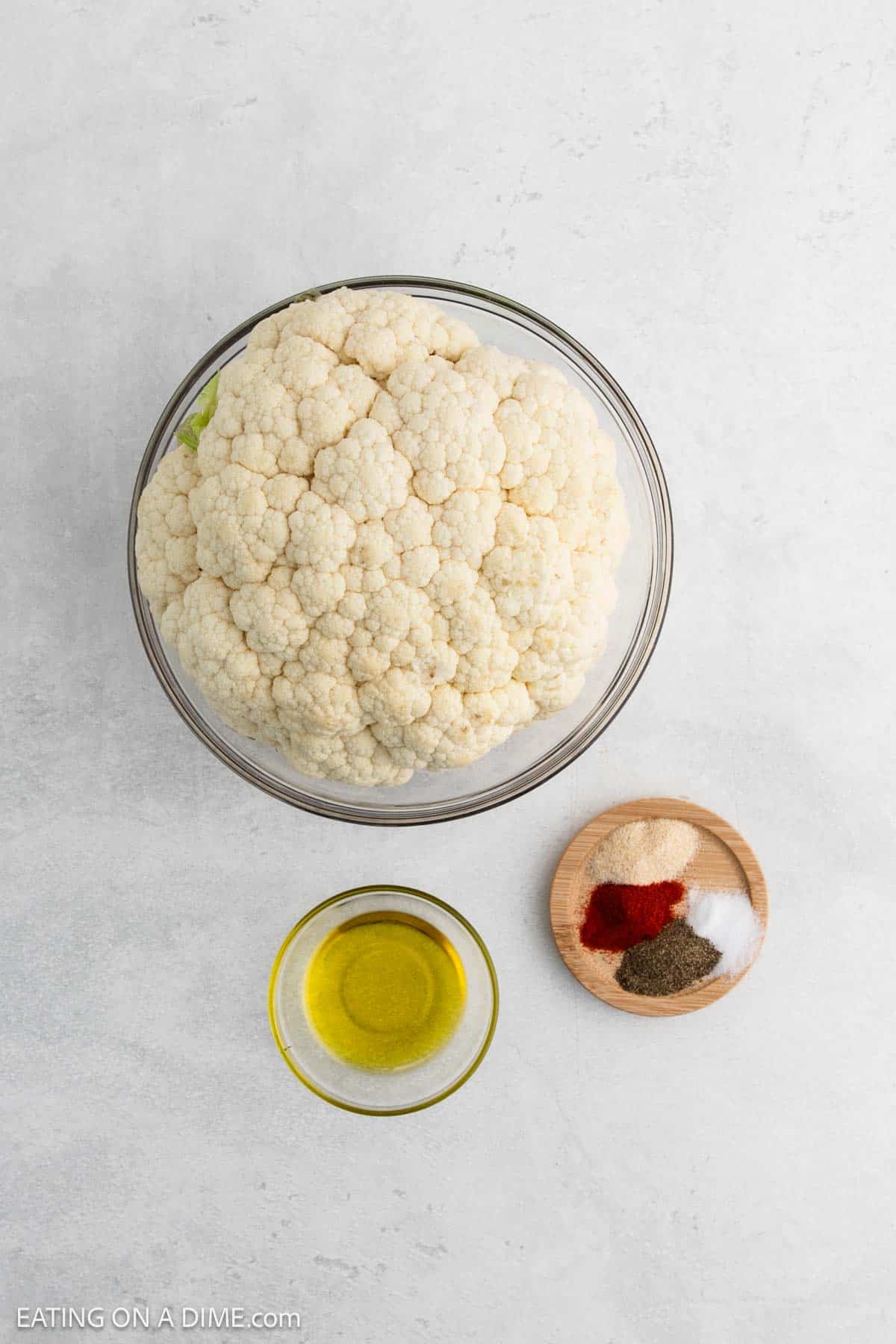 A large bowl of whole cauliflower is ready to become roasted cauliflower perfection, sitting next to a small glass bowl of olive oil and a wooden dish filled with assorted spices like red pepper, black pepper, salt, and garlic powder on a light gray surface.