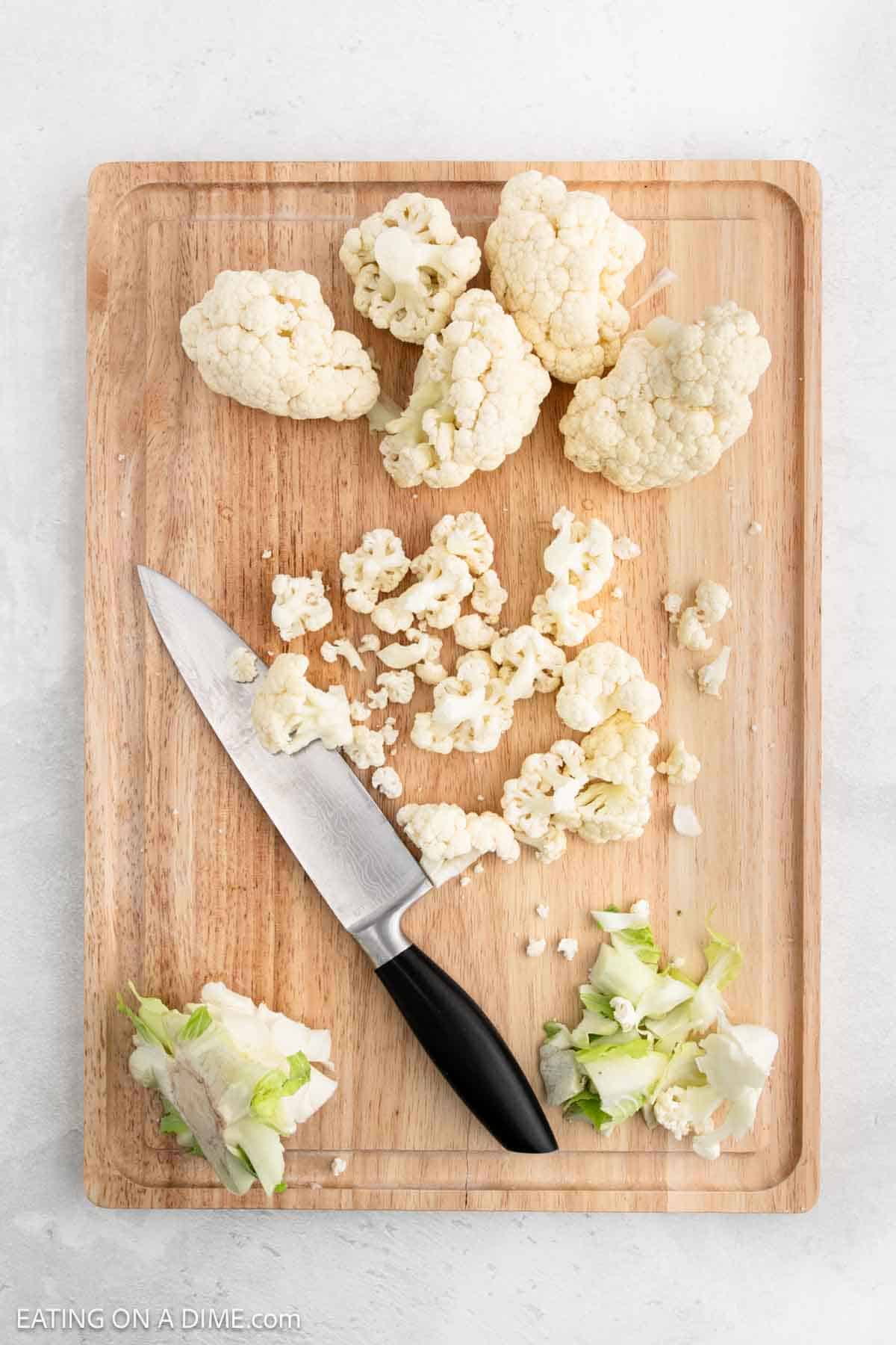 Chopped cauliflower florets and leaves rest on a wooden cutting board with a kitchen knife, ready to transform into delicious roasted cauliflower.