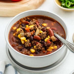 A hearty bowl of chili filled with ground beef, kidney beans, corn, diced tomatoes, and onions resembles a taco soup. A spoon rests inside on a white surface with a small dish of sliced jalapeños and part of a tomato visible in the background.