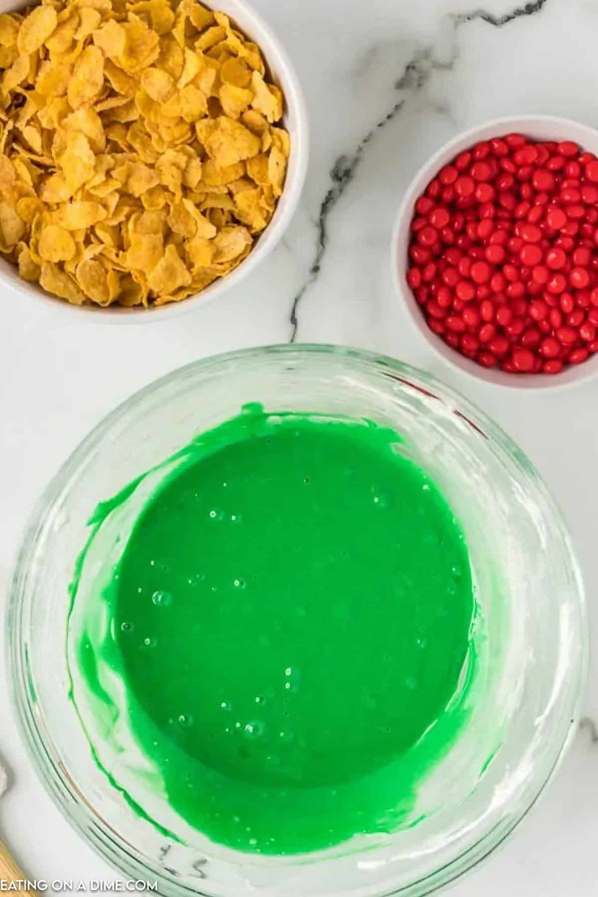 Green melted marshmallow mixture in a bowl with a spatula, with a bowl of corn flakes on the side with a small bowl of red candy on the side