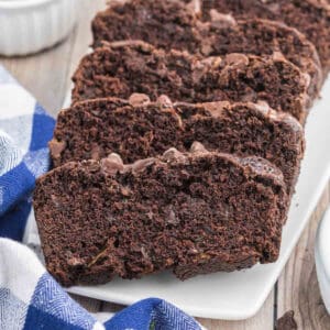 Sliced chocolate zucchini bread elegantly arranged on a white platter. The moist, rich slices are studded with visible chocolate chips. A blue and white checkered cloth peeks out from the side, adding a touch of color to the rustic presentation of this delectable treat.