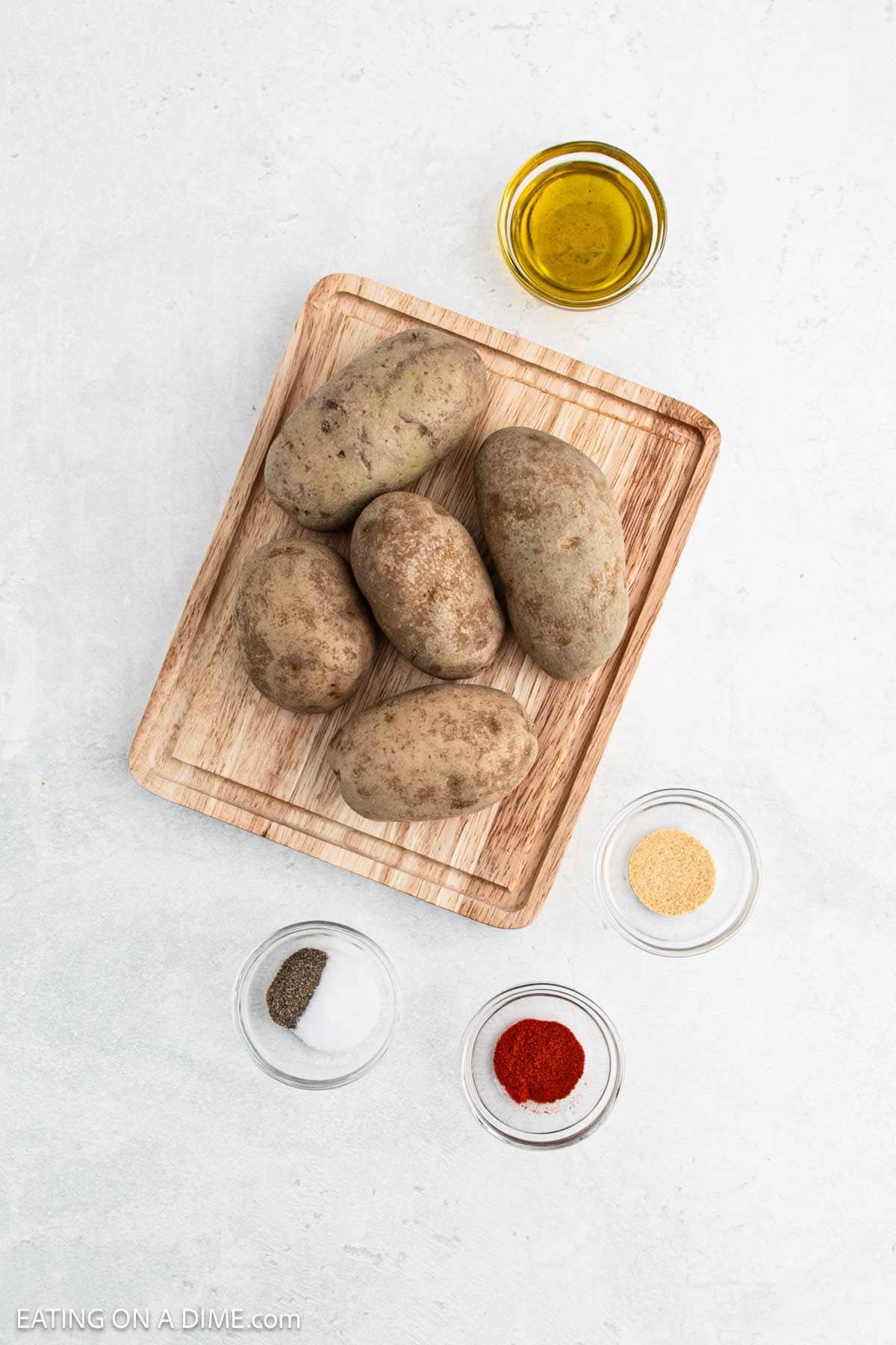 Wooden tray with five unpeeled potatoes, reminiscent of the perfect start to homemade French fries, surrounded by small bowls containing olive oil, garlic powder, paprika, salt, and pepper on a light surface.