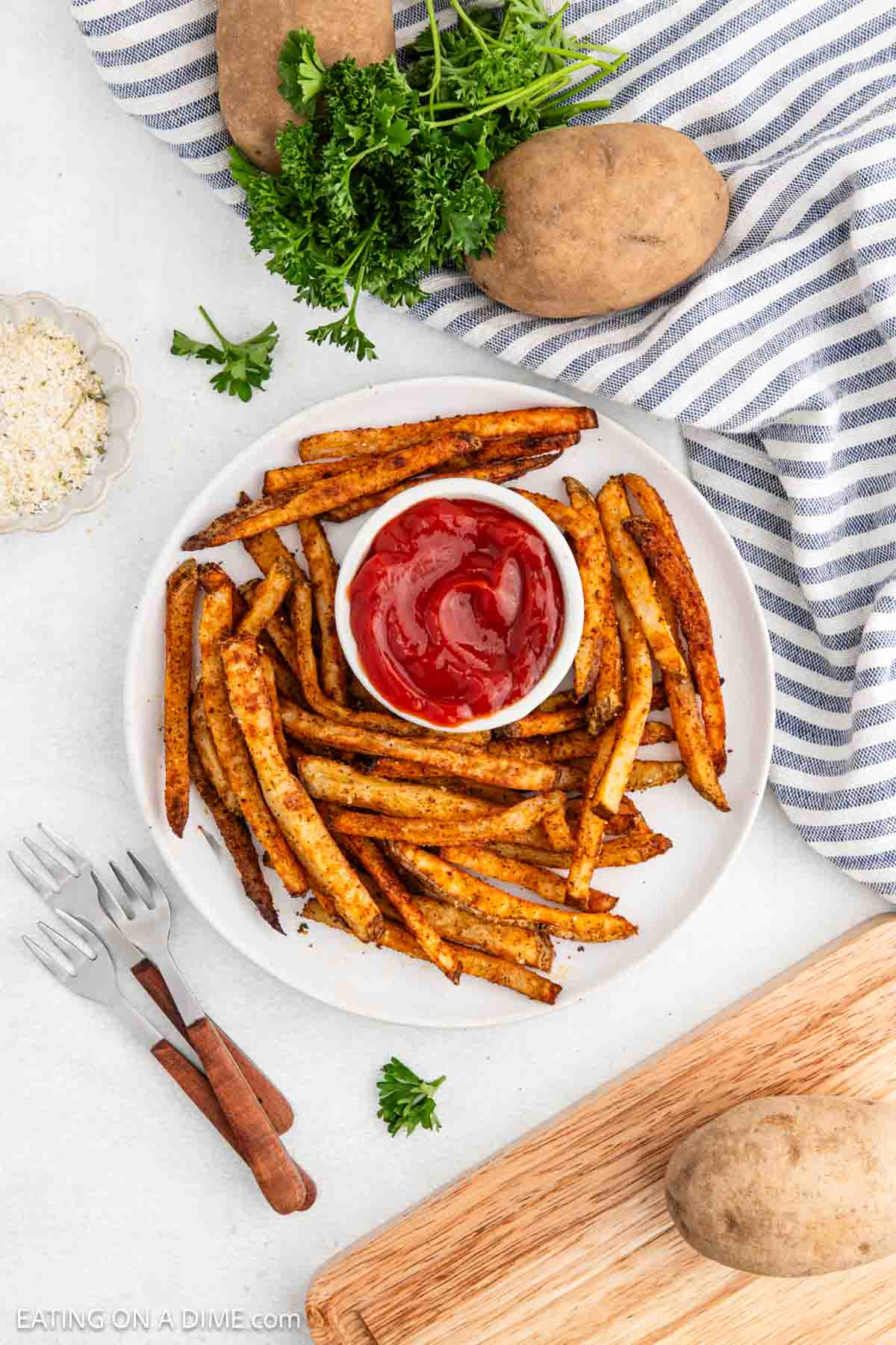 A plate of seasoned baked French fries surrounds a small dish of ketchup. Fresh potatoes, parsley, and a striped cloth are nearby. Small forks, grated cheese, and a wooden board with a potato add to the setting.