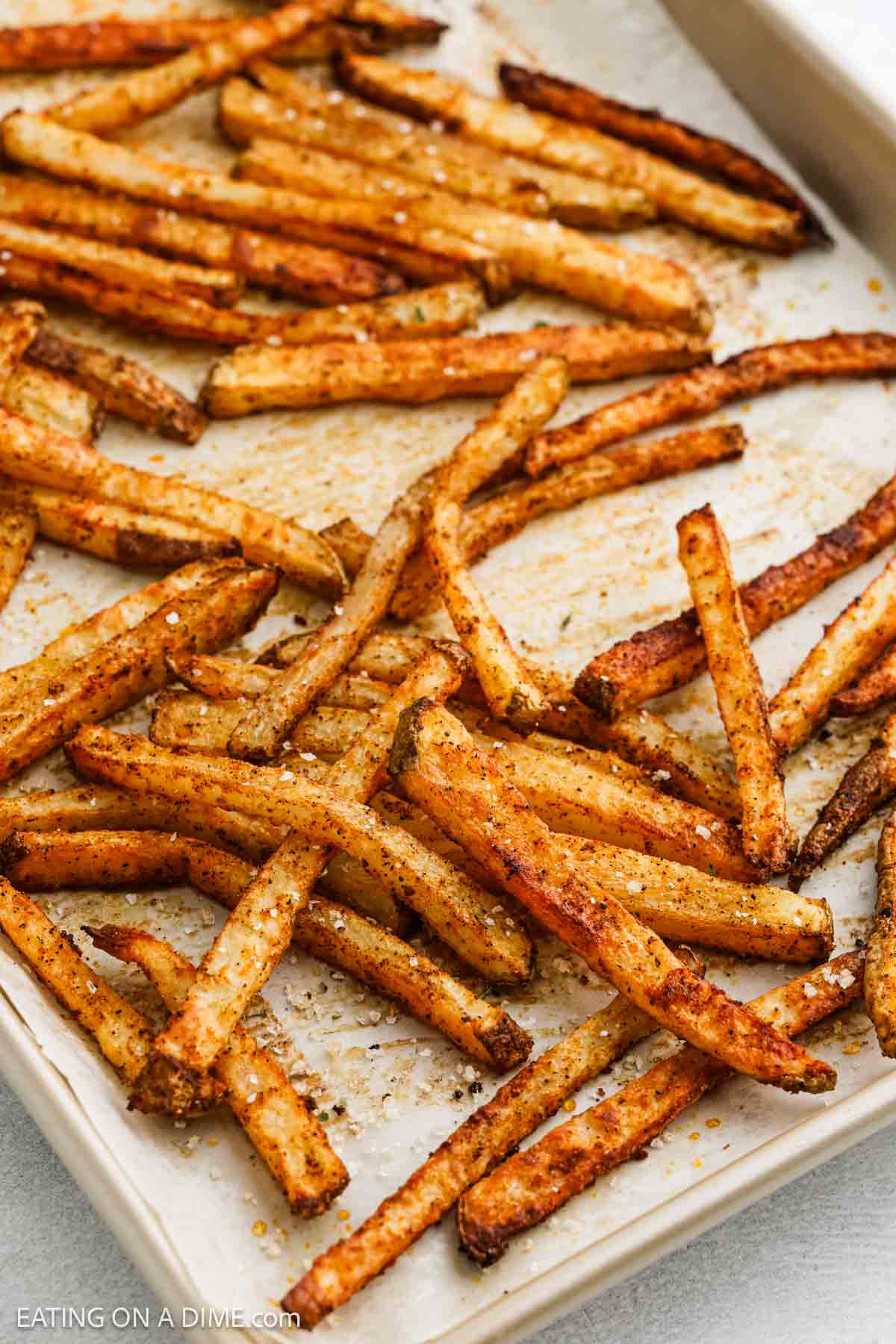 A baking sheet brimming with crispy, golden-brown baked French fries lies scattered and freshly pulled from the oven. The fries boast a visible sprinkle of black pepper and spices, enhancing their well-seasoned allure.