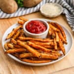 A plate of baked French fries surrounds a small bowl of ketchup on a wooden table. In the background, there are potatoes and a small dish with creamy sauce.