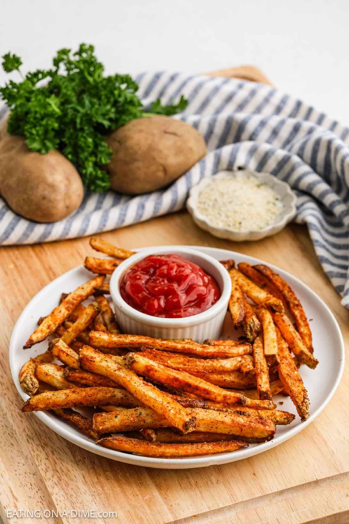 A plate of crispy baked French fries is served with a small dish of ketchup in the center. In the background, two whole potatoes, a bunch of parsley, and a bowl of seasoning rest on a wooden surface beside a striped cloth.