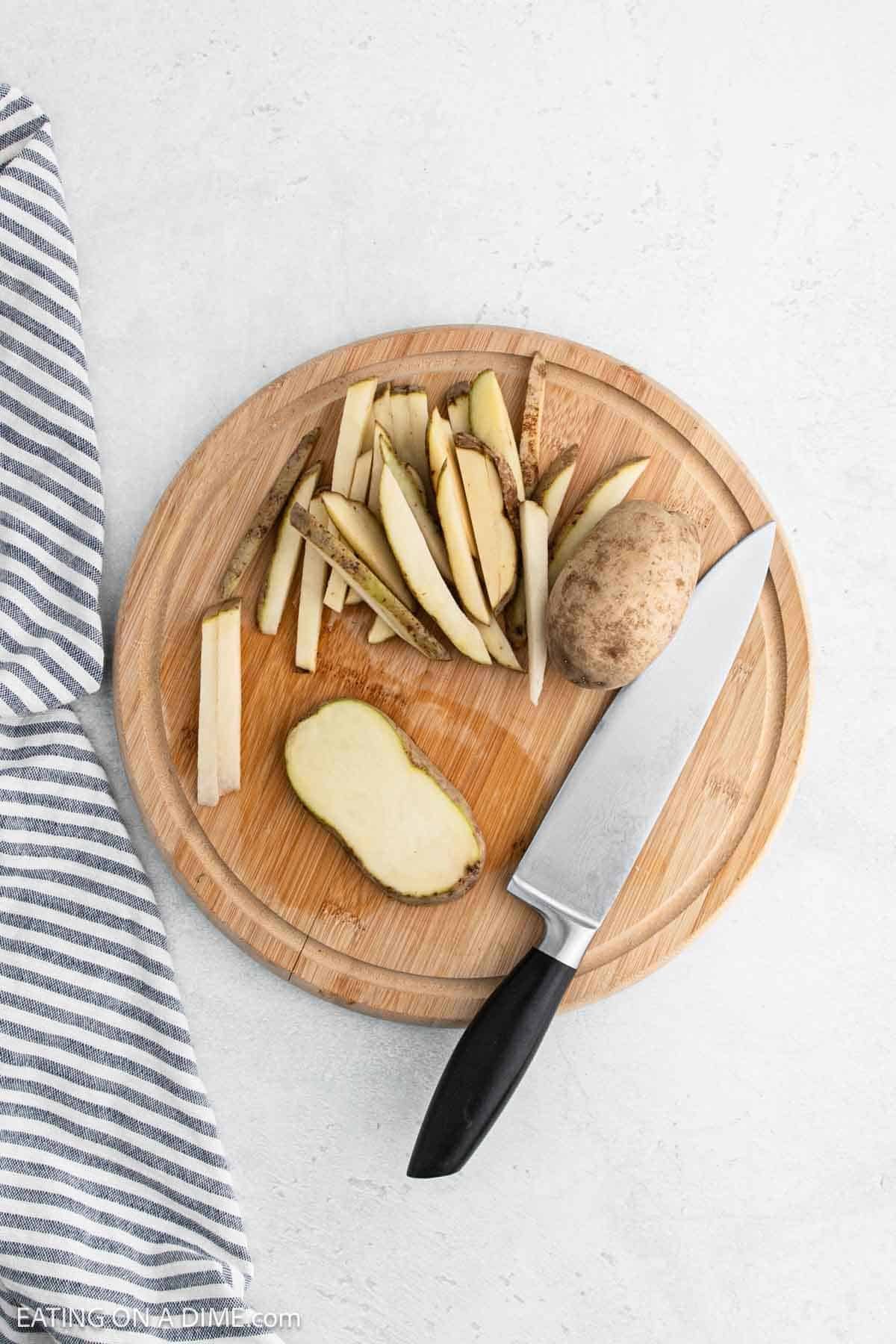 A circular wooden cutting board holds sliced potatoes ready for baking, alongside an unsliced potato and a knife. A striped cloth peeks from the left side, set against a light, textured backdrop perfect for preparing delicious homemade French fries.