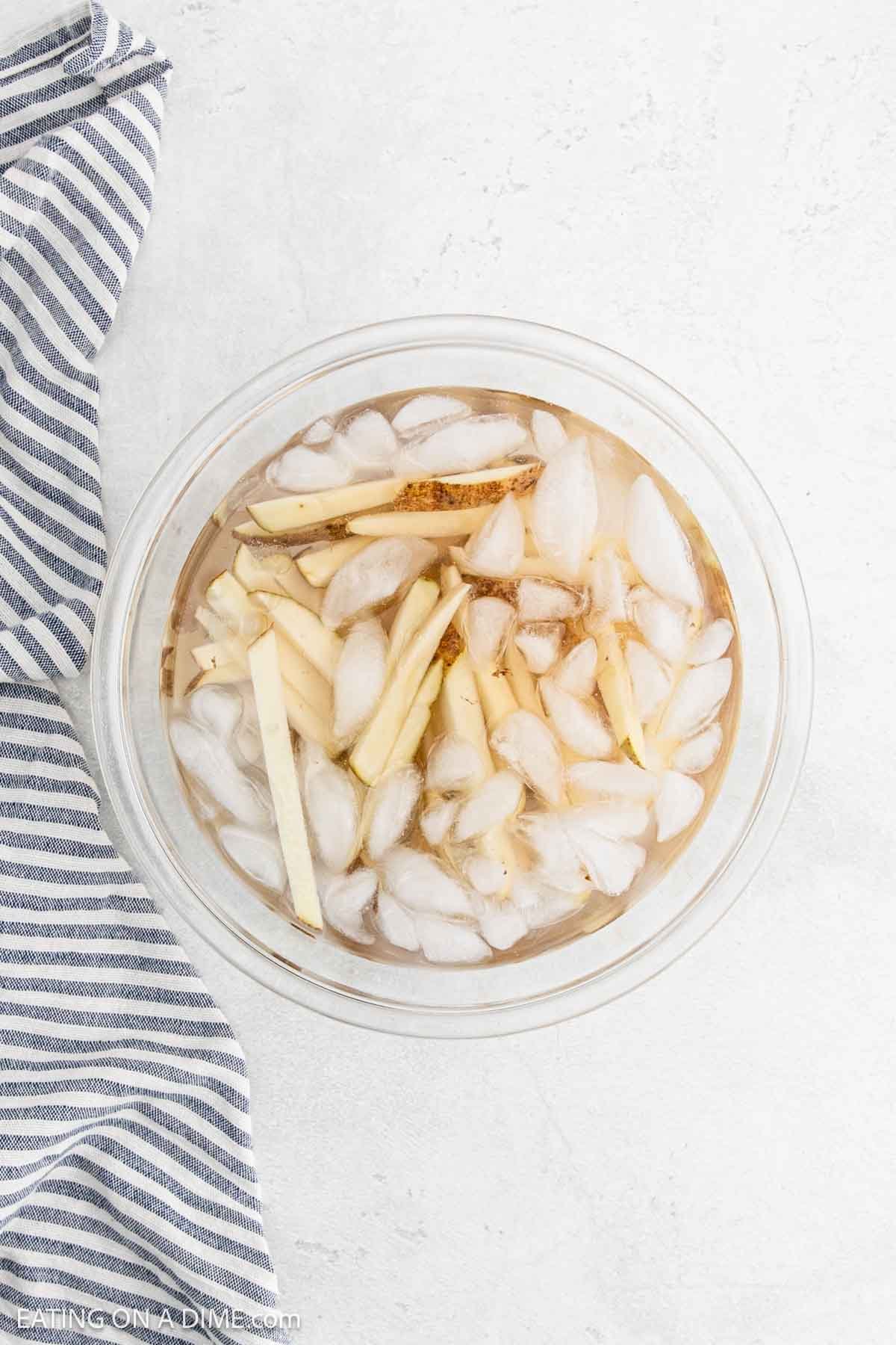 A top view reveals a bowl of sliced potatoes, destined to become crispy French fries, soaking with ice cubes. The bowl rests on a light surface beside a striped cloth.