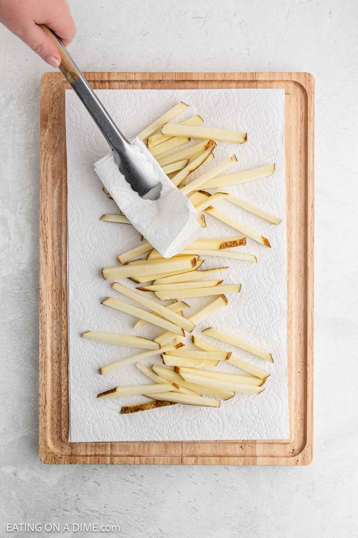 A hand uses tongs to place raw potato strips on a paper towel-covered wooden cutting board. The potatoes are being dried, likely in preparation for baking French fries, on a light gray surface.