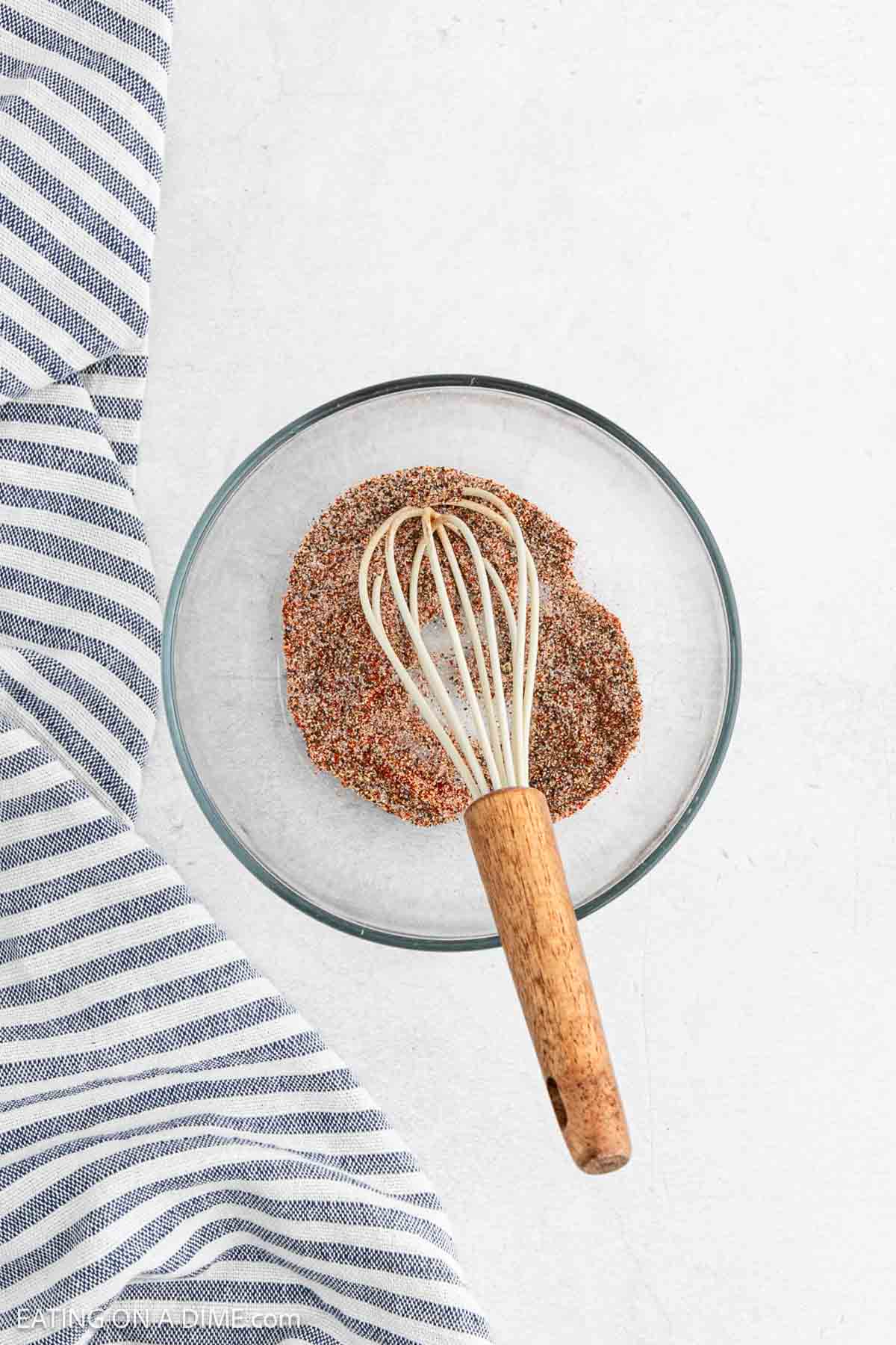 A glass bowl filled with a spice mixture and a whisk inside, resting on a white surface. A blue and white striped cloth is partially visible on the left, perfect for seasoning baked French fries with flair.