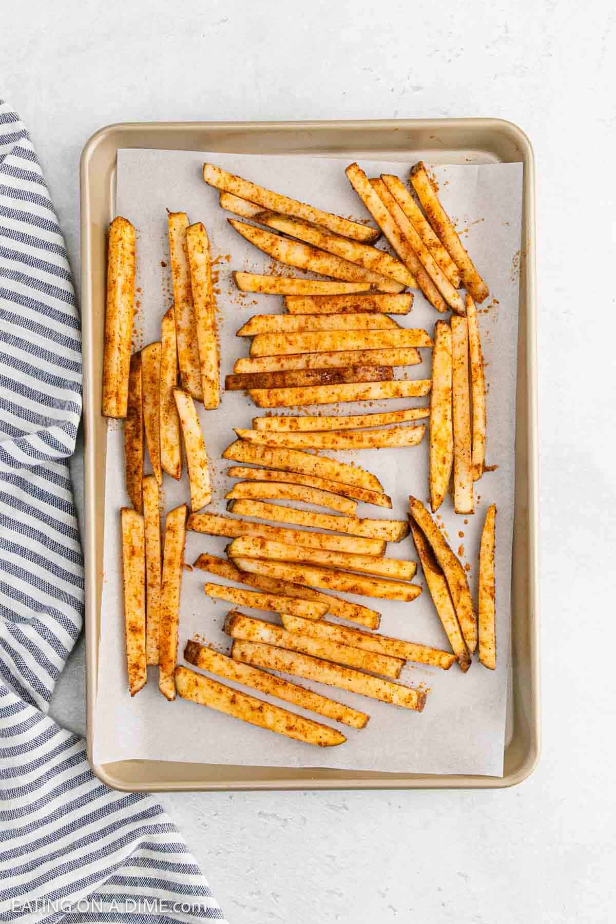 Baked potato fries seasoned with spices are spread out on a parchment-covered baking sheet. The baking sheet is placed on a light surface next to a gray and white striped cloth, evoking the classic allure of French fries with a healthier twist.