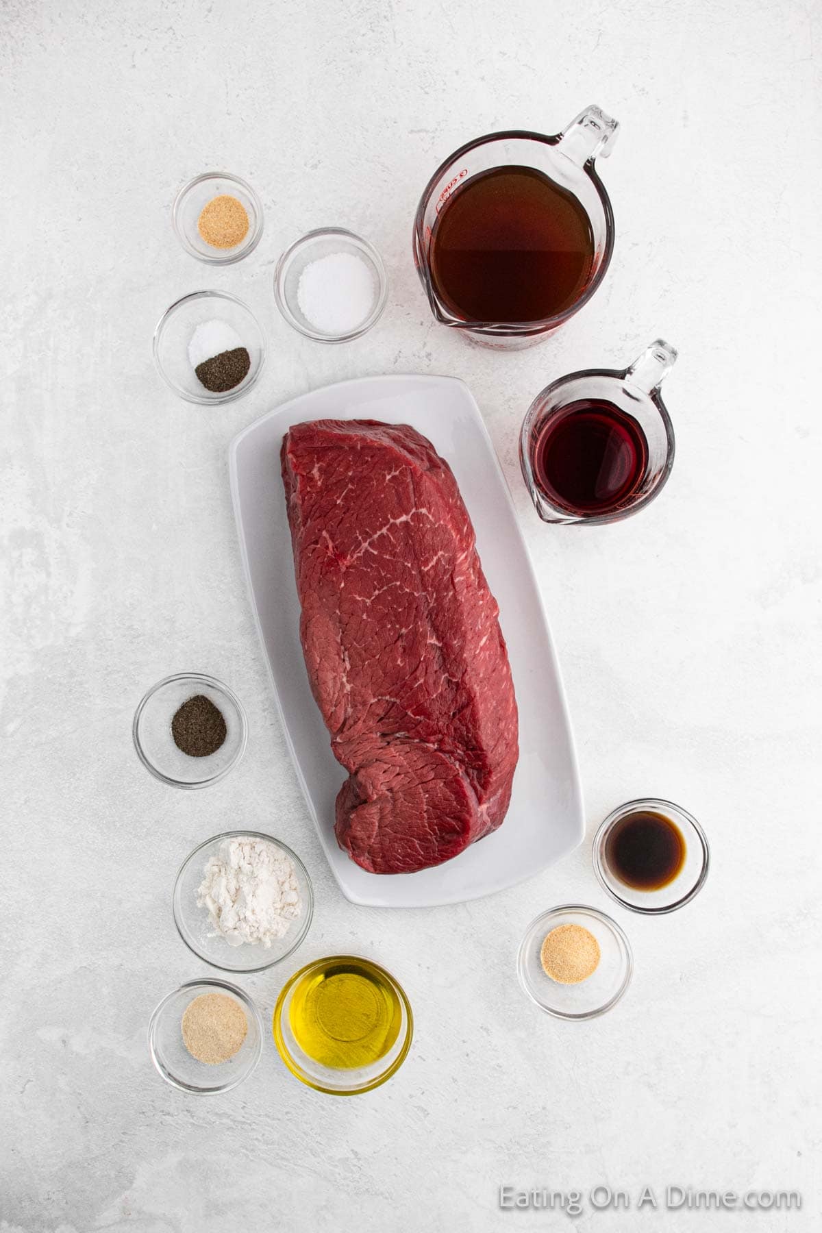 A raw beef roast on a rectangular white plate, surrounded by small bowls and measuring cups containing various ingredients: broth, red wine, soy sauce, olive oil, and spices, set against a light background.