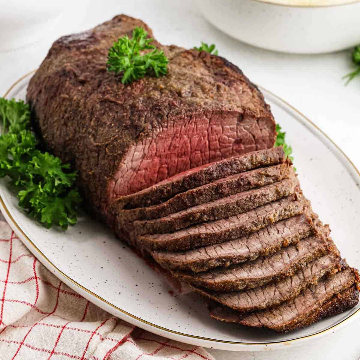 Sliced roast beef on a white oval platter, garnished with fresh parsley. The meat is cooked medium, showing a juicy pink center, and is served with a red and white checkered cloth beside the plate.