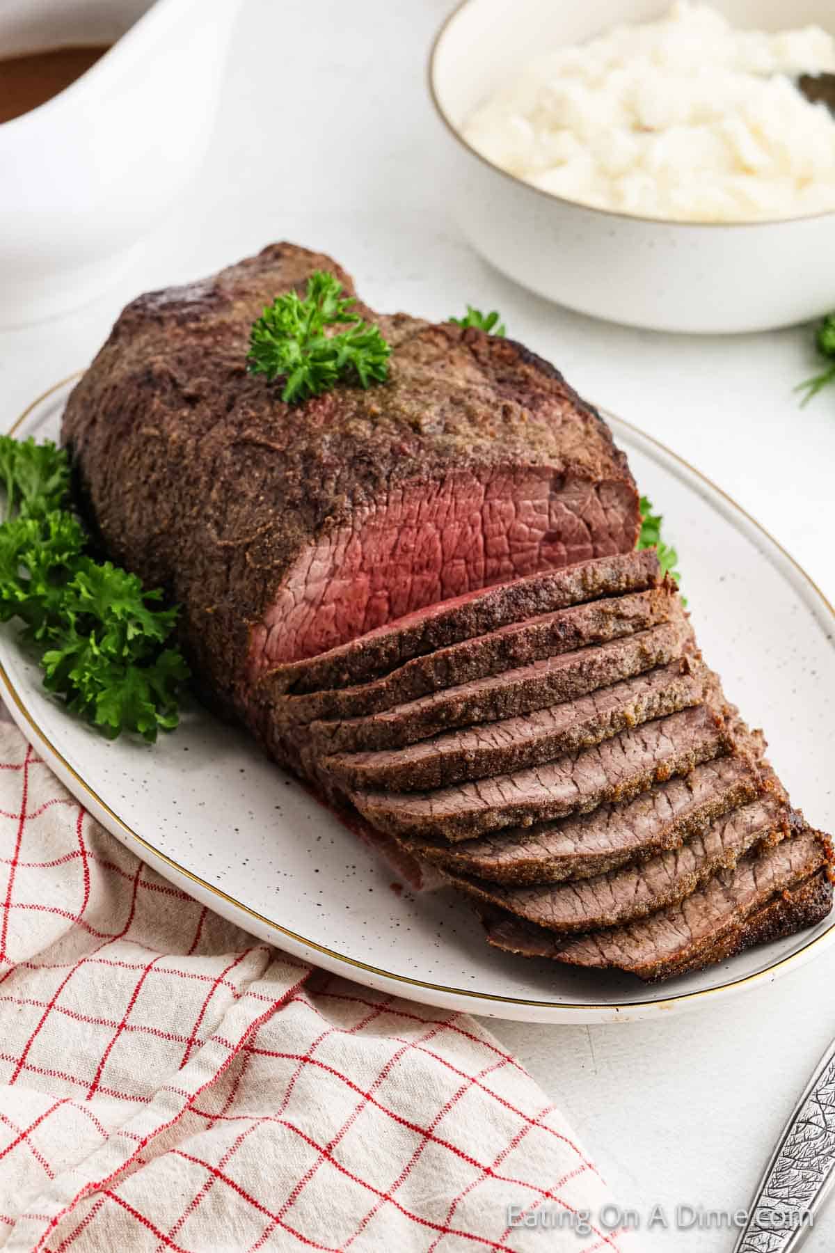 Sliced roast beef garnished with parsley on a white oval plate, placed on a table with a red-checkered cloth. In the background, there's a bowl of mashed potatoes and a gravy boat.