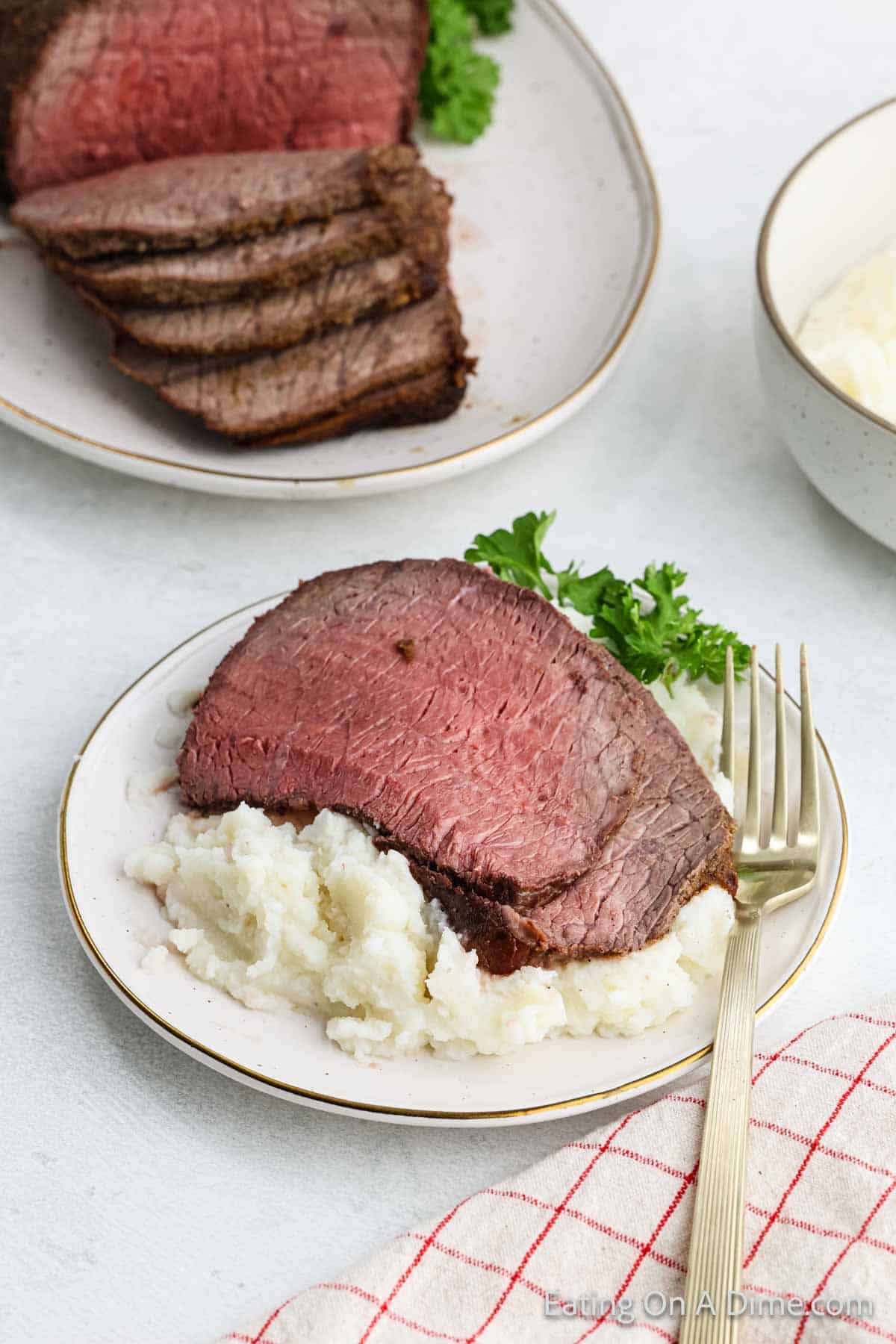 Slices of roast beef served over mashed potatoes on a white plate with a sprig of parsley. A fork rests on the side. In the background, more sliced roast beef is displayed on another plate. A red-checkered napkin is nearby.