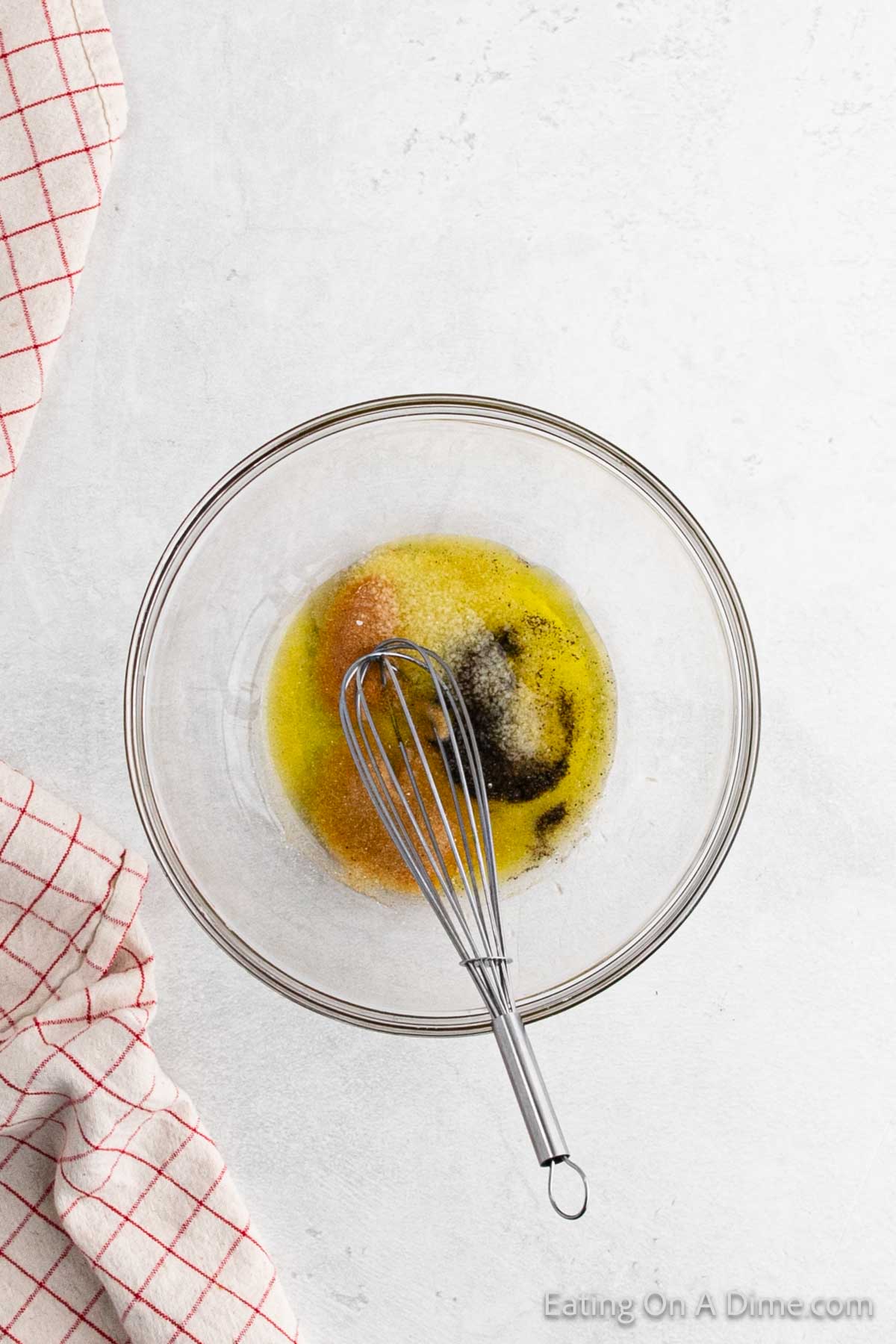 A clear glass bowl contains brown sugar, melted butter, and a whisk on a white surface. A red and white checkered kitchen towel is partially visible on the left.