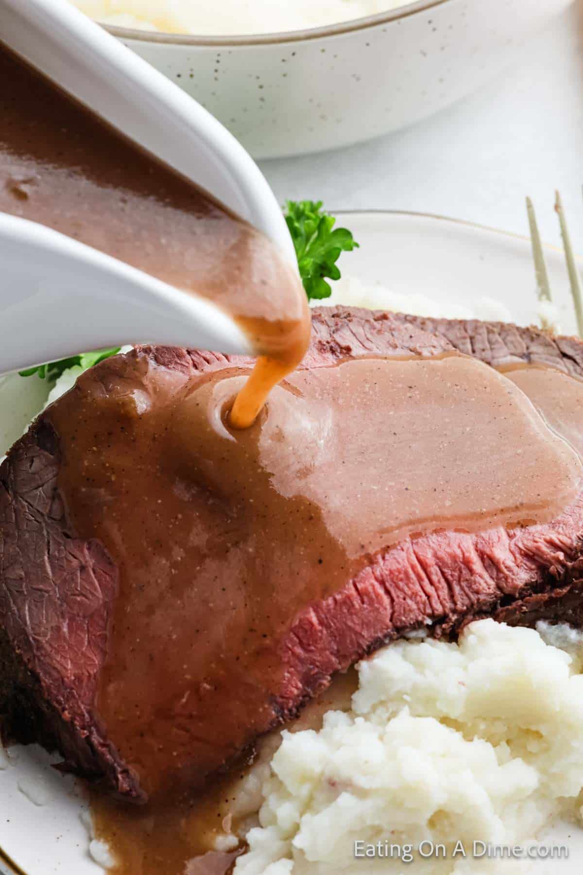 Close-up of gravy being poured from a white gravy boat over a thick slice of roast beef, accompanied by a serving of creamy mashed potatoes. A sprig of parsley adds a touch of garnish on the plate.