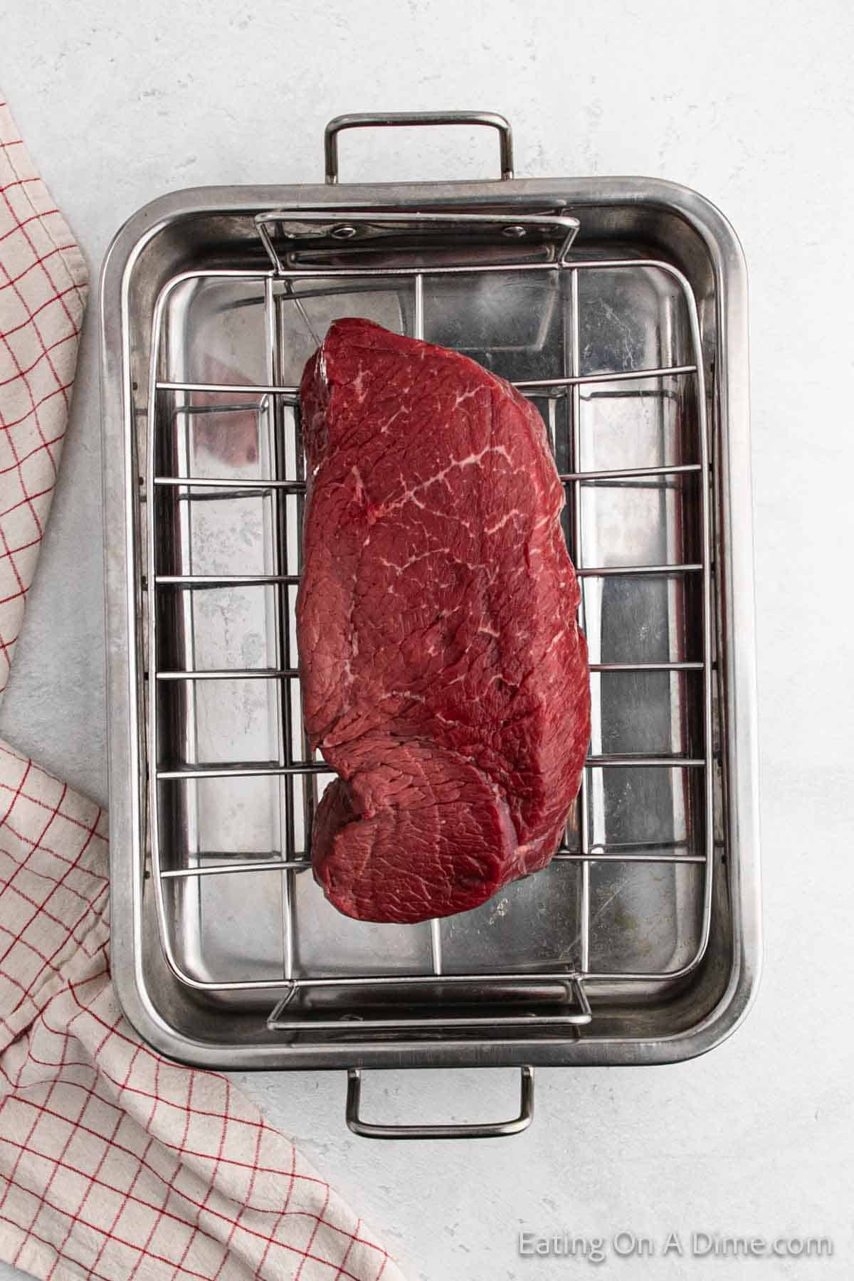 A raw beef roast sits on a metal rack inside a roasting pan. A checkered kitchen towel is placed beside the pan on the light-colored surface.