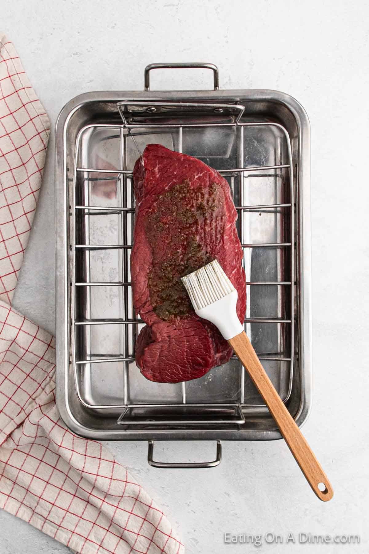 A raw beef roast in a metal roasting pan on a rack. A wooden-handled brush resting on the meat applies marinade. A red and white checkered cloth is partially visible on the side.