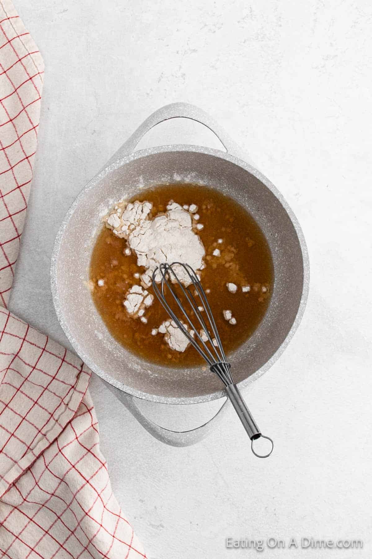 A pot containing a mixture of brown liquid and flour with a whisk resting inside. The pot is on a white surface next to a red-checkered cloth.