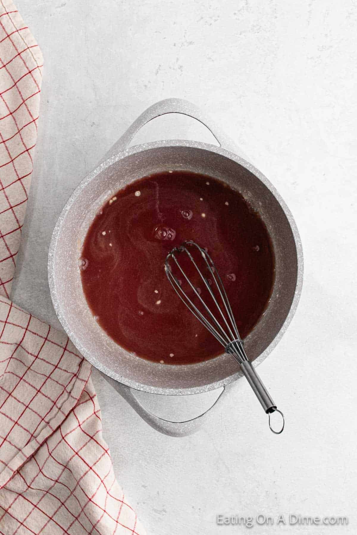 A pot of cranberry sauce being whisked, placed on a white countertop next to a red and white checkered kitchen towel.