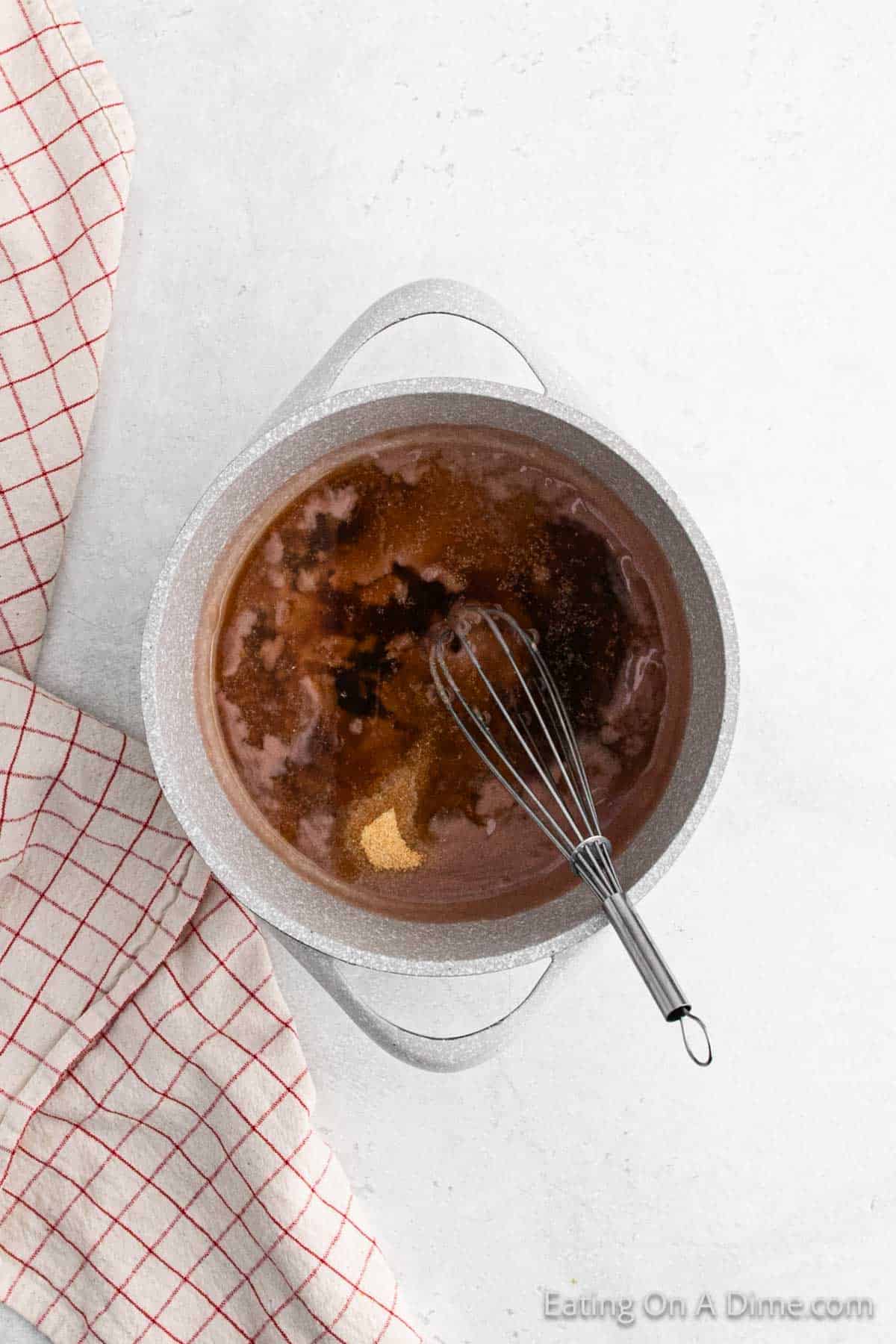 A pot with melted chocolate and a whisk inside is placed on a light-colored surface. A checked kitchen towel with red lines is partially visible on the left side.