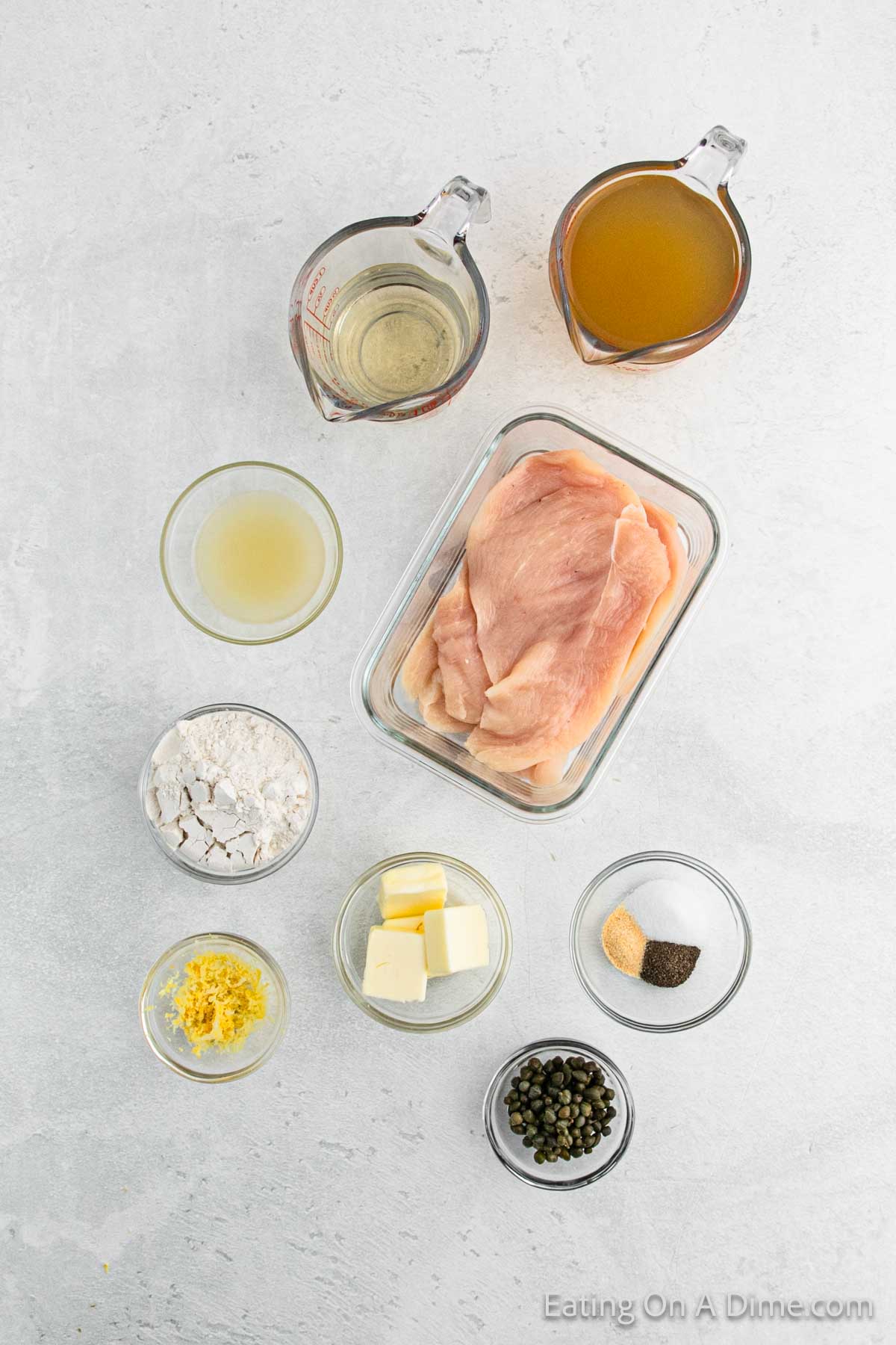 Ingredients for Chicken Piccata are displayed on a light surface: raw chicken breasts, flour, butter, lemon juice, lemon zest, capers, seasoning, chicken broth, and white wine in various glass containers.