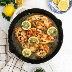 A pan of cooked Chicken Piccata garnished with lemon slices and capers sits on a light surface. Fresh parsley and lemon halves are nearby, along with a blue and white plate containing more lemon slices. A striped kitchen towel is in view, enhancing this delightful chicken recipe.