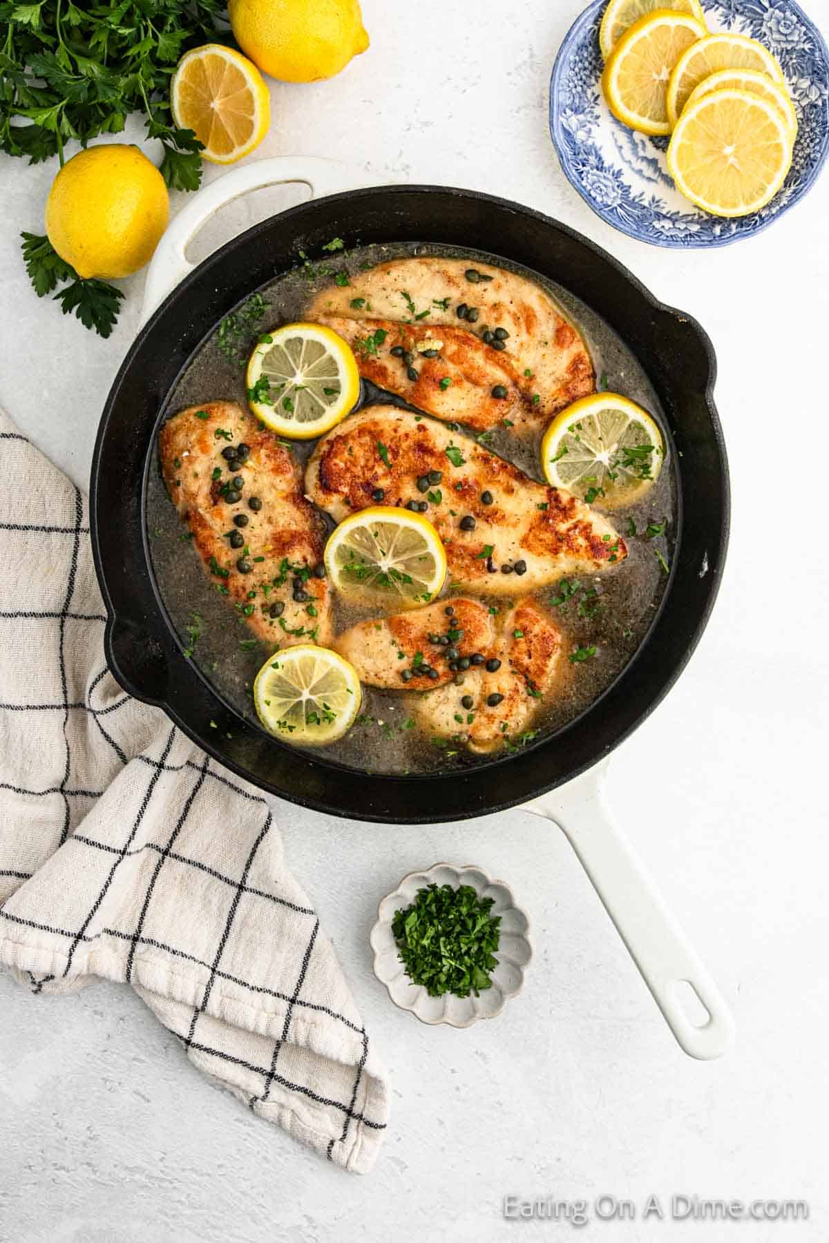 A skillet with Chicken Piccata in a lemon-caper sauce, garnished with lemon slices and chopped herbs. Nearby are fresh lemons, a blue plate with lemon slices, a checkered cloth, and a small dish of chopped herbs—perfect for your next recipe.