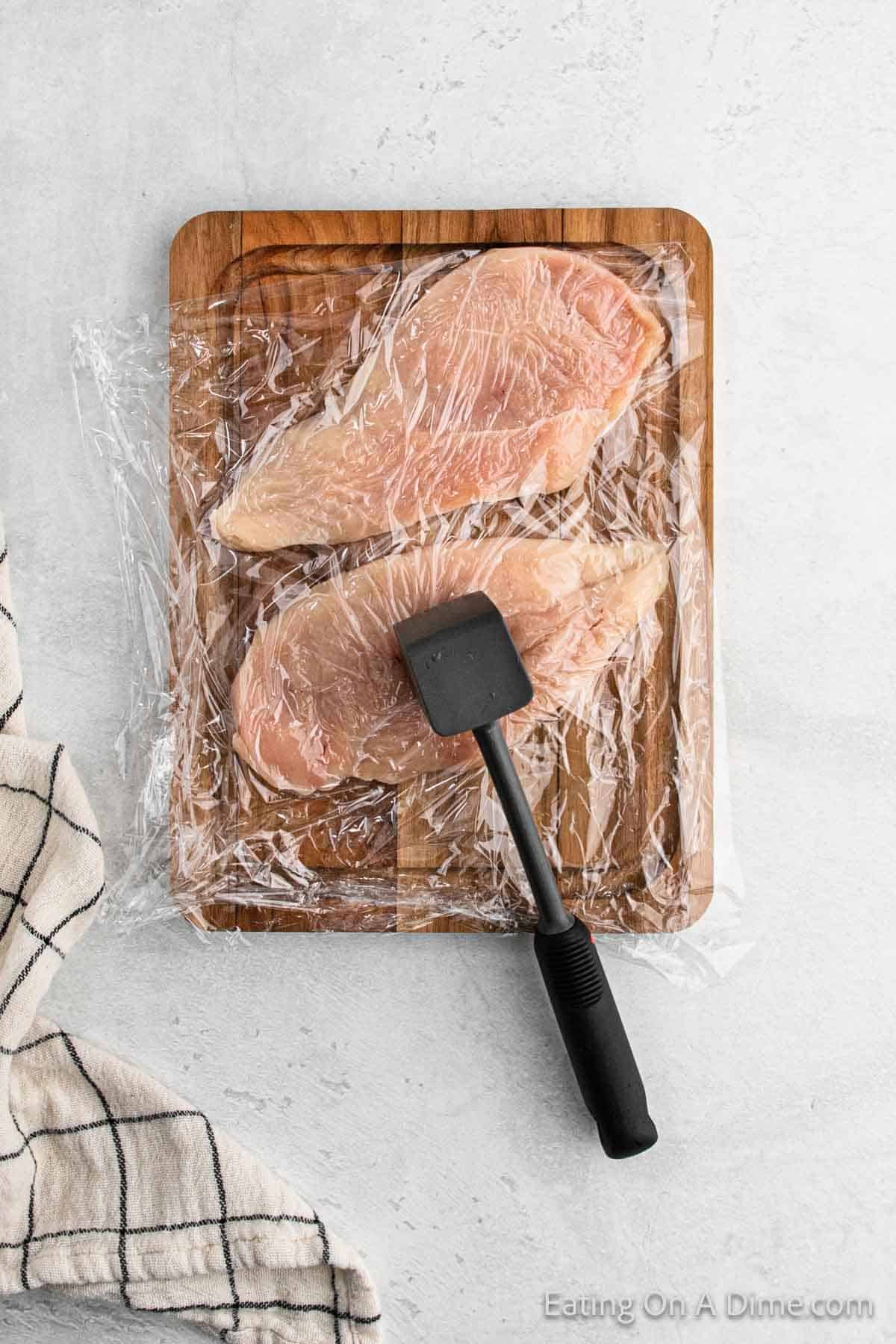 Two raw chicken breasts, perfect for a classic Chicken Piccata, are covered with plastic wrap on a wooden cutting board, with a meat tenderizer resting on top. A white and black checkered cloth is partly visible on the left side, setting the stage for this delicious Italian cuisine preparation.