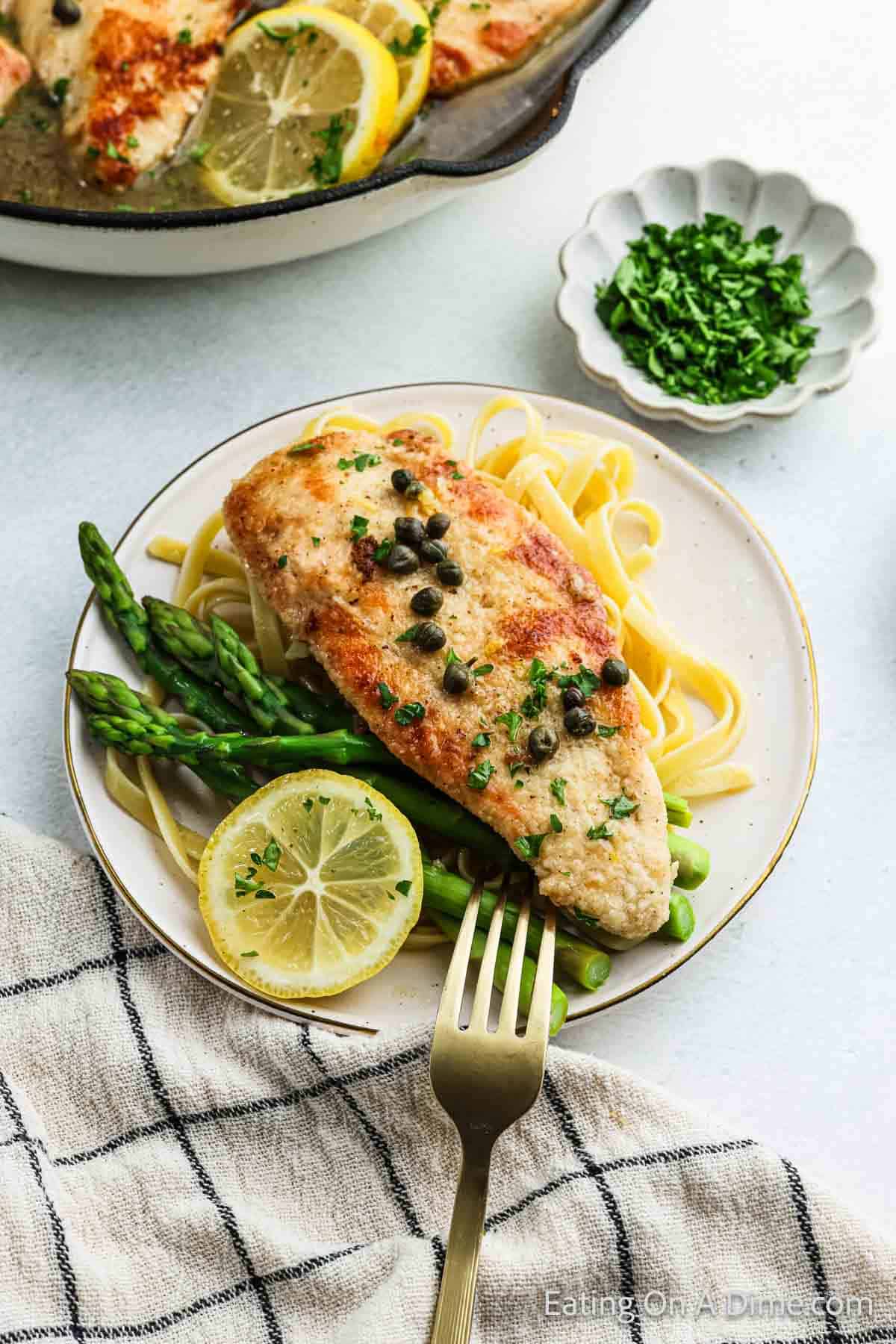 A plate with Chicken Piccata, garnished with capers and parsley, sits atop a bed of fettuccine and asparagus. Lemon slices accompany this classic Italian cuisine dish. A fork rests on the plate, while a bowl of chopped herbs is nearby.