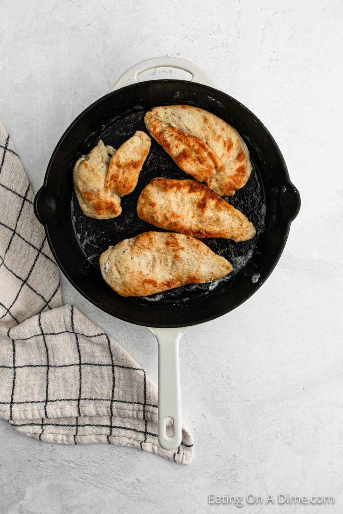 A cast iron skillet with four cooked chicken breasts, lightly browned and seasoned, reminiscent of Chicken Piccata, on a gray surface. A white and black checkered cloth is beside the skillet.