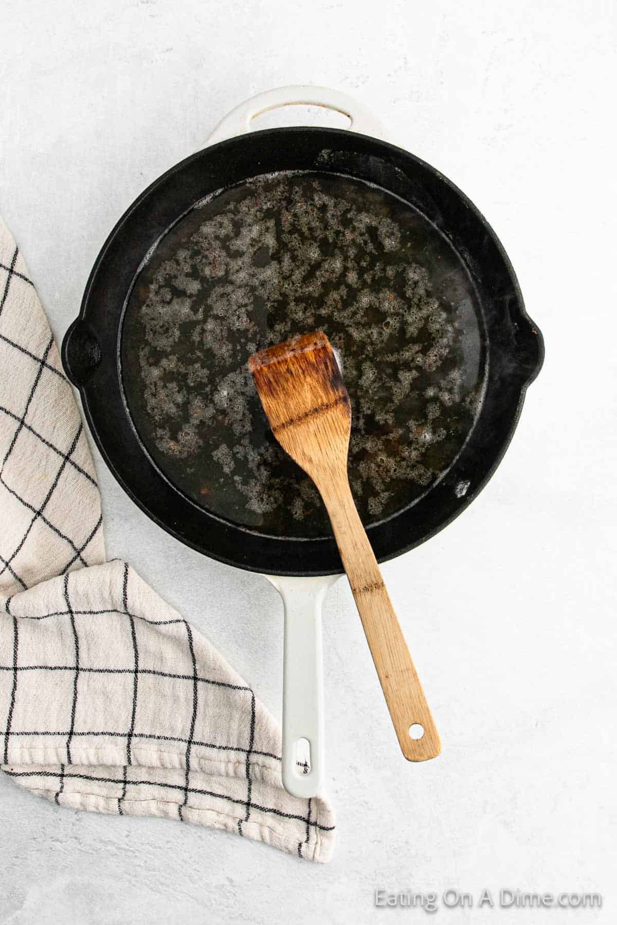 A white pan with a black interior rests on a light surface, suggesting the aftermath of a delicious Chicken Piccata. Inside, a wooden spatula lies atop some food residue. To the left, a beige cloth with a black grid pattern partially peeks into view.