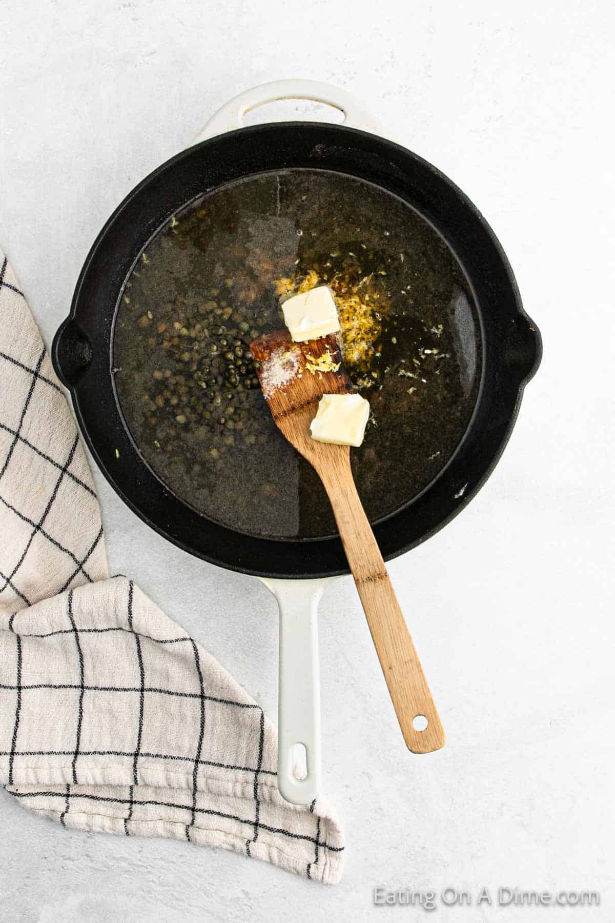 A white pan contains lentils simmering in broth with three pats of butter and a wooden spatula, evoking the comforting essence of a chicken piccata dish. A checkered kitchen towel is draped beside the pan on a light surface.