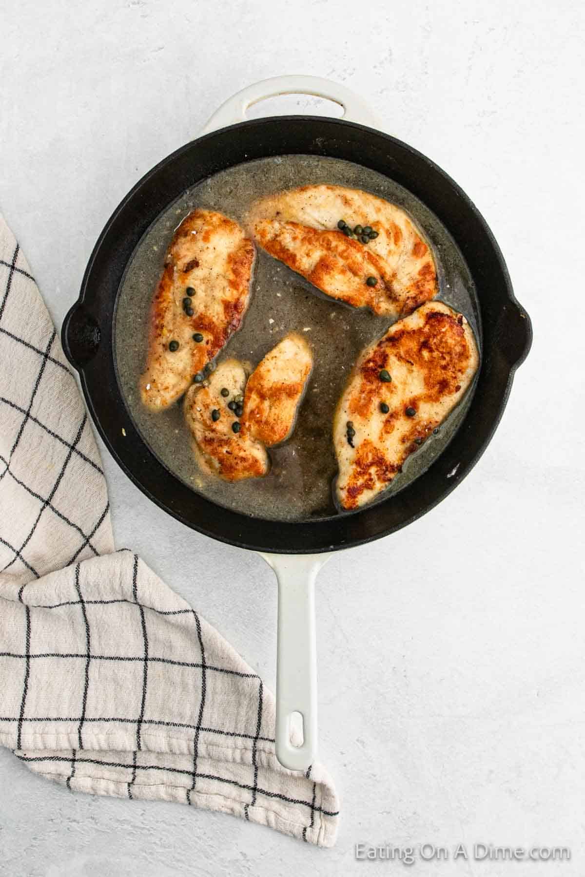 A skillet with Chicken Piccata featuring succulent chicken breasts in a creamy sauce, garnished with capers. A white and black checkered napkin is placed beside the skillet on a light gray surface.