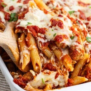 A close-up of a baked penne pasta dish, part of Meal Plan 39, with melted cheese and tomato sauce. A wooden spoon lifts a portion from the casserole dish, revealing layers of pasta, sauce, and garnished herbs on top.