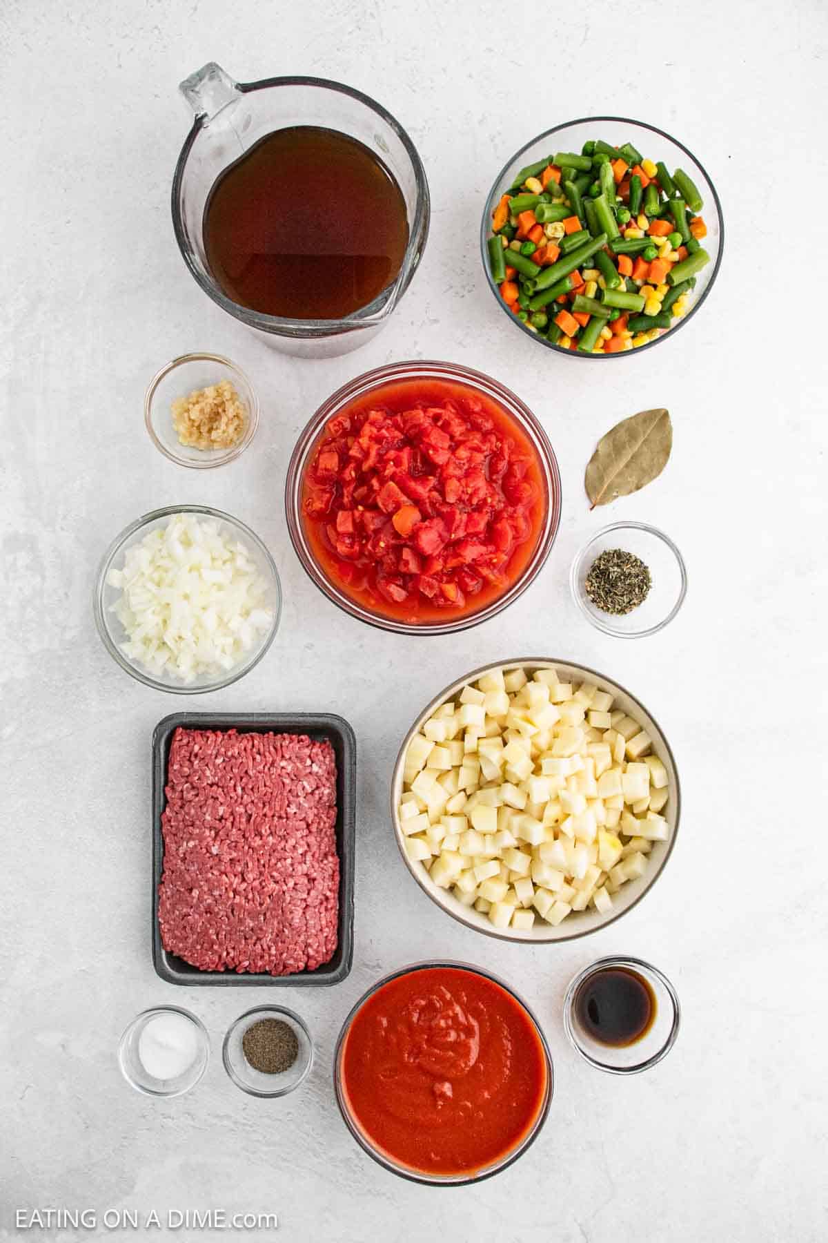 Ingredients for a comforting hamburger soup laid out on a white surface: beef broth in a measuring cup, mixed vegetables, minced garlic, diced tomatoes, bay leaf, Italian seasoning, chopped onion, raw beef, potato cubes, tomato sauce, pepper, and Worcestershire sauce.