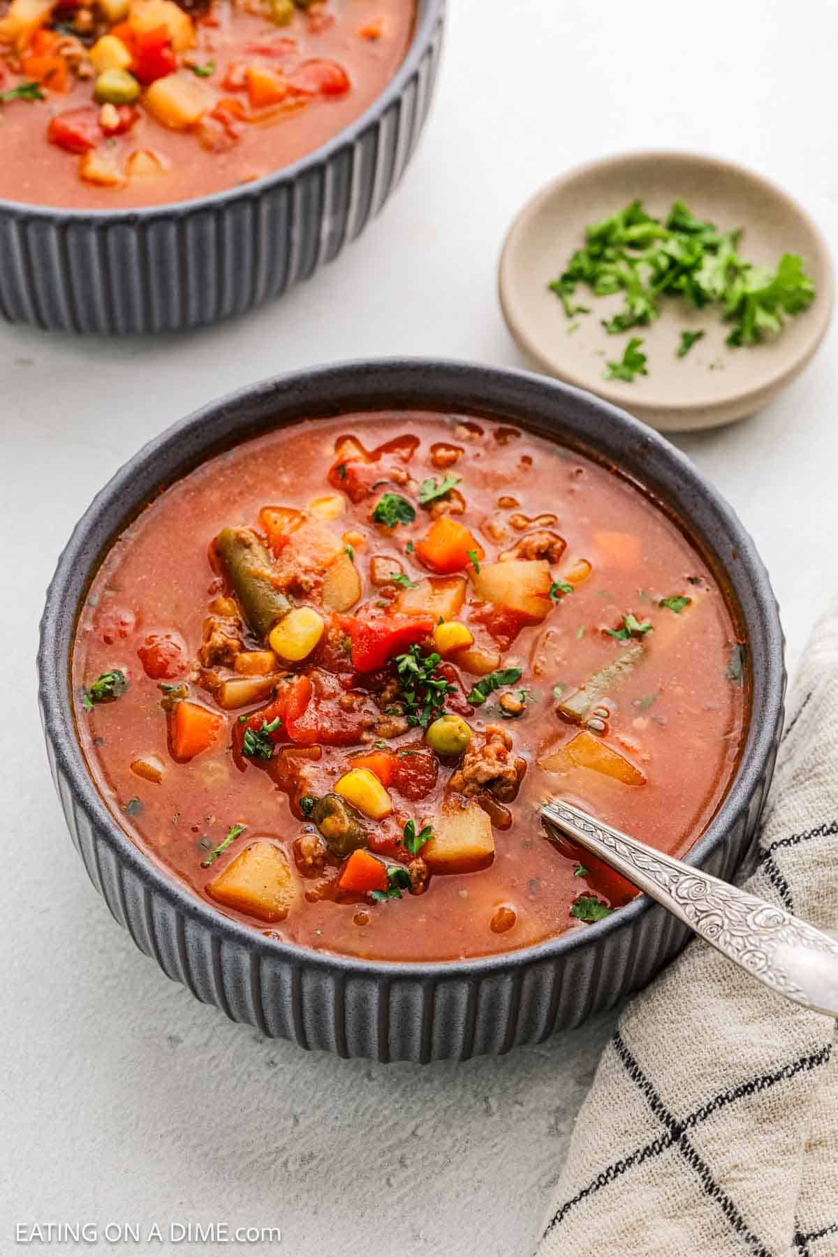 A vegetable soup recipe featuring chunks of potatoes, peas, corn, carrots, and green beans in a tomato-based broth. The soup is garnished with chopped herbs and accompanied by a small dish of additional herbs and a striped napkin.